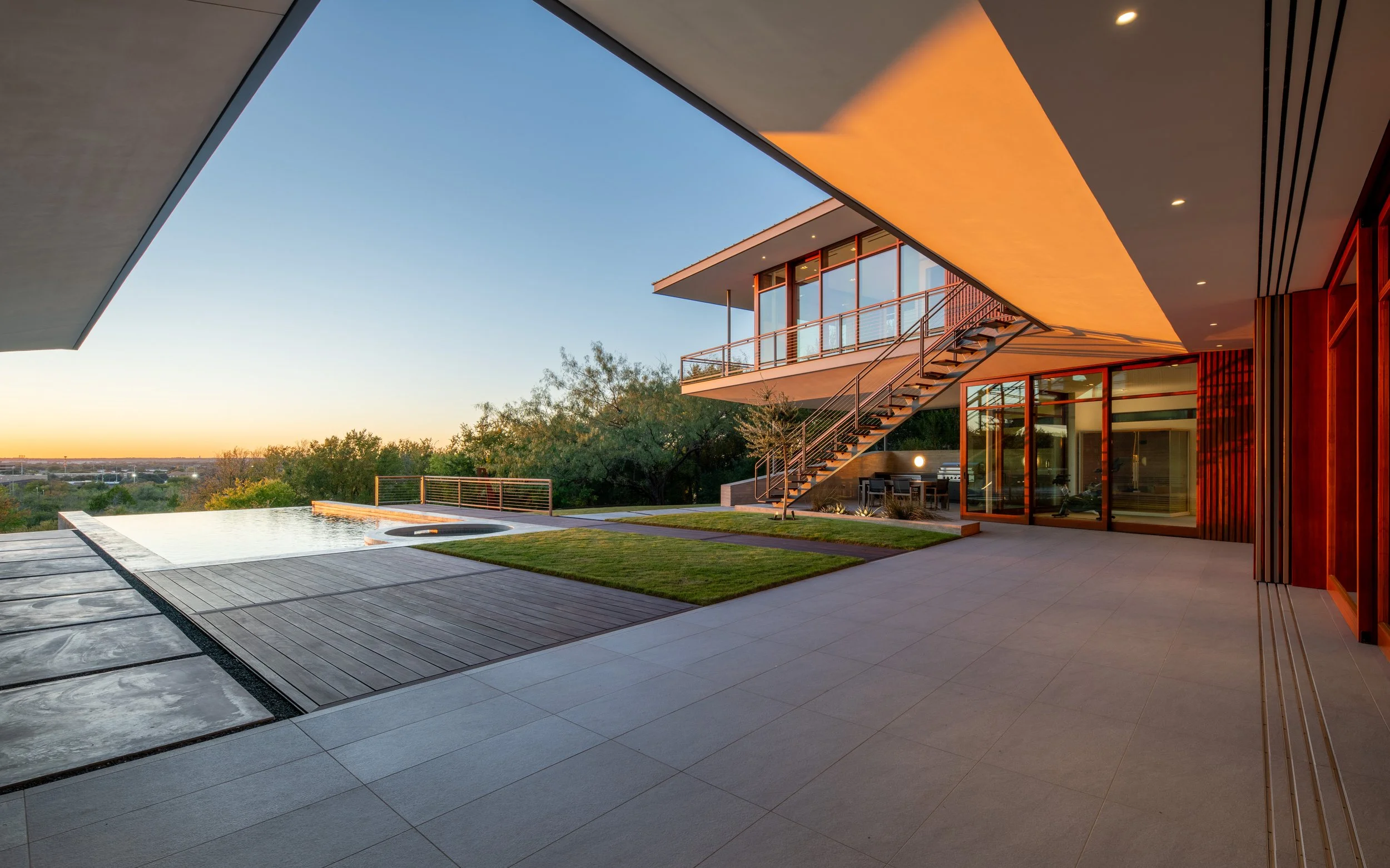 Modern house with outdoor terrace, infinity pool, grassy area, and stairway, overlooking a sunset and distant landscape.