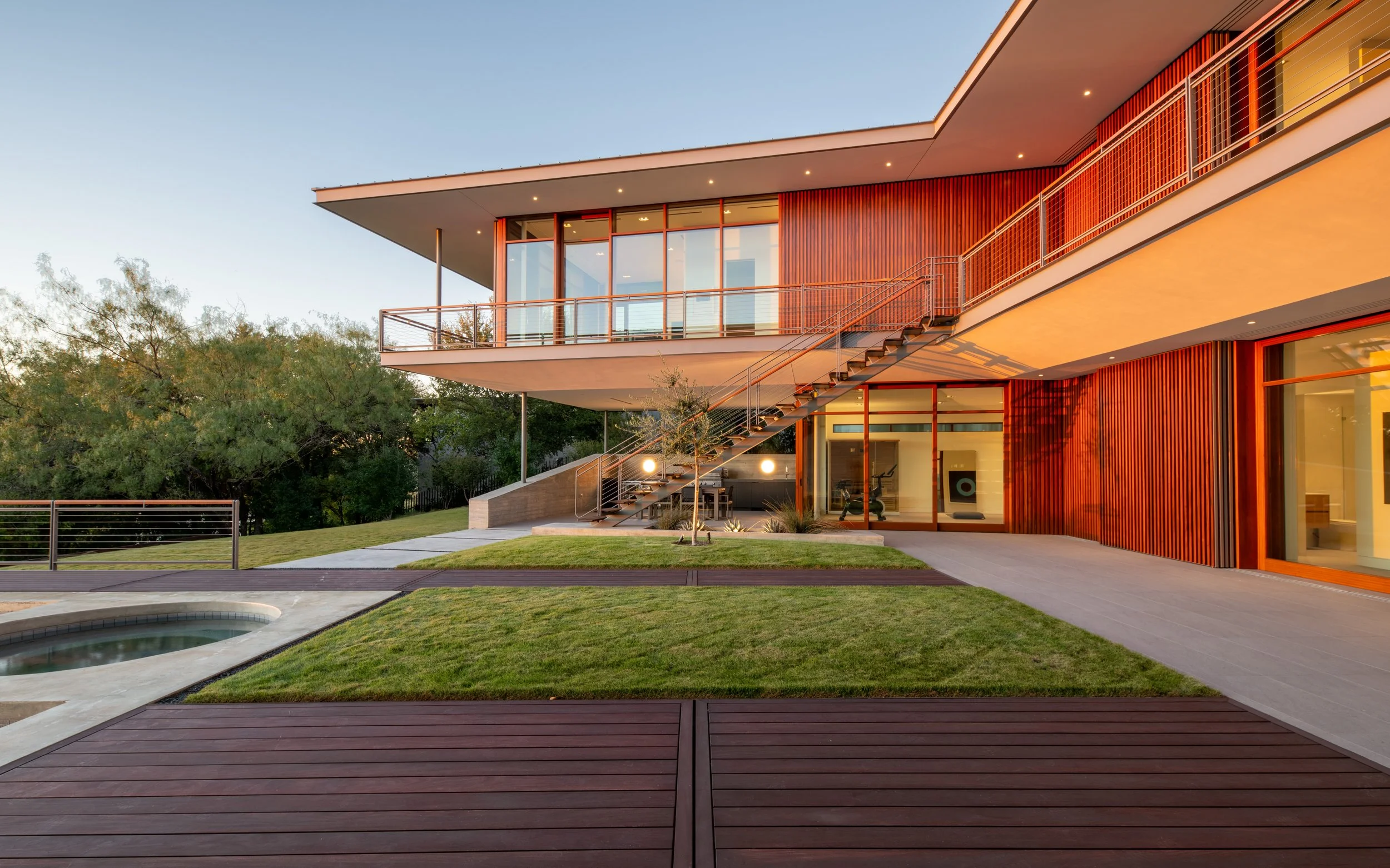 Modern two-story house with large glass windows, wooden accent walls, and outdoor stairs leading to a deck, surrounded by a landscaped yard with grass, trees, and a small pool.