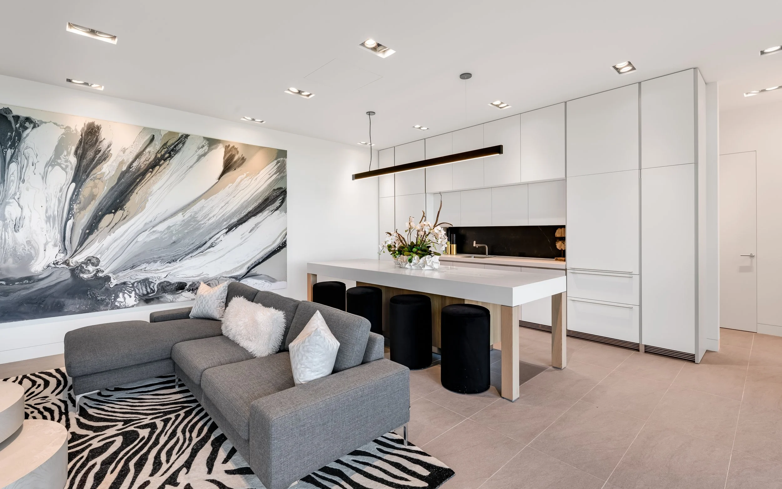 Modern open-concept living room and kitchen with a gray sectional sofa, decorative pillows, a large abstract black and white wall art, a white dining counter with black stools, minimalist white cabinets, and a flower arrangement on the island.