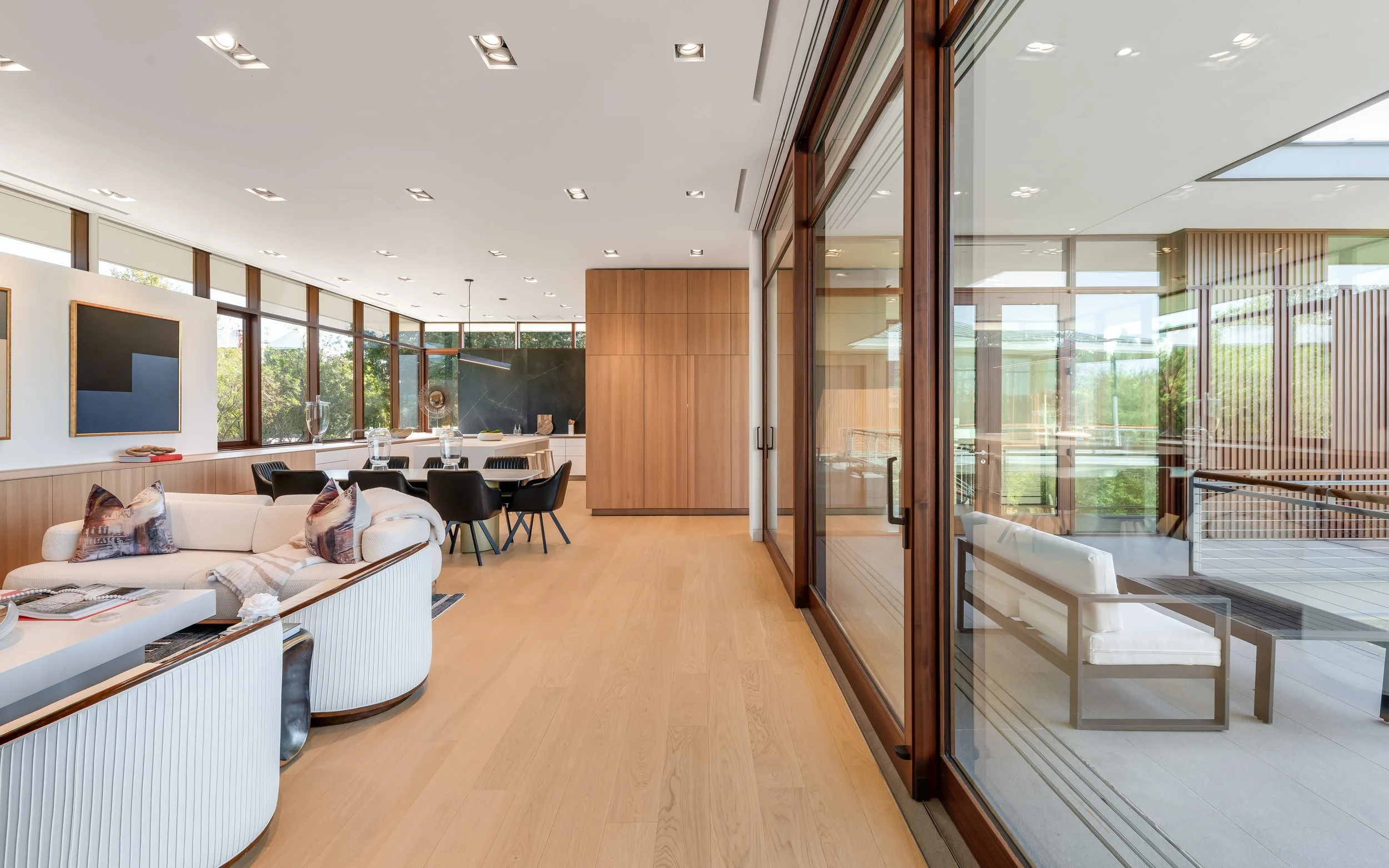 Modern living room with large glass windows, wooden floors, white sofa with pillows, black dining chairs around a table, and a mix of minimalist decor.