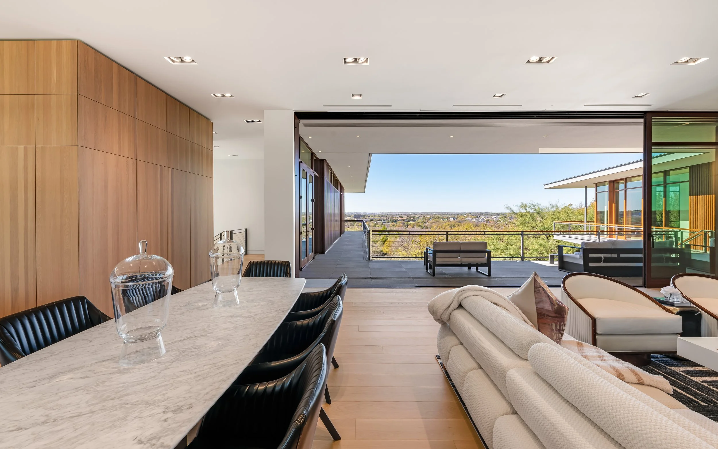 Modern living room with a view of a balcony, wooden wall, marble dining table with chairs, and a seating area with a sofa and armchairs.