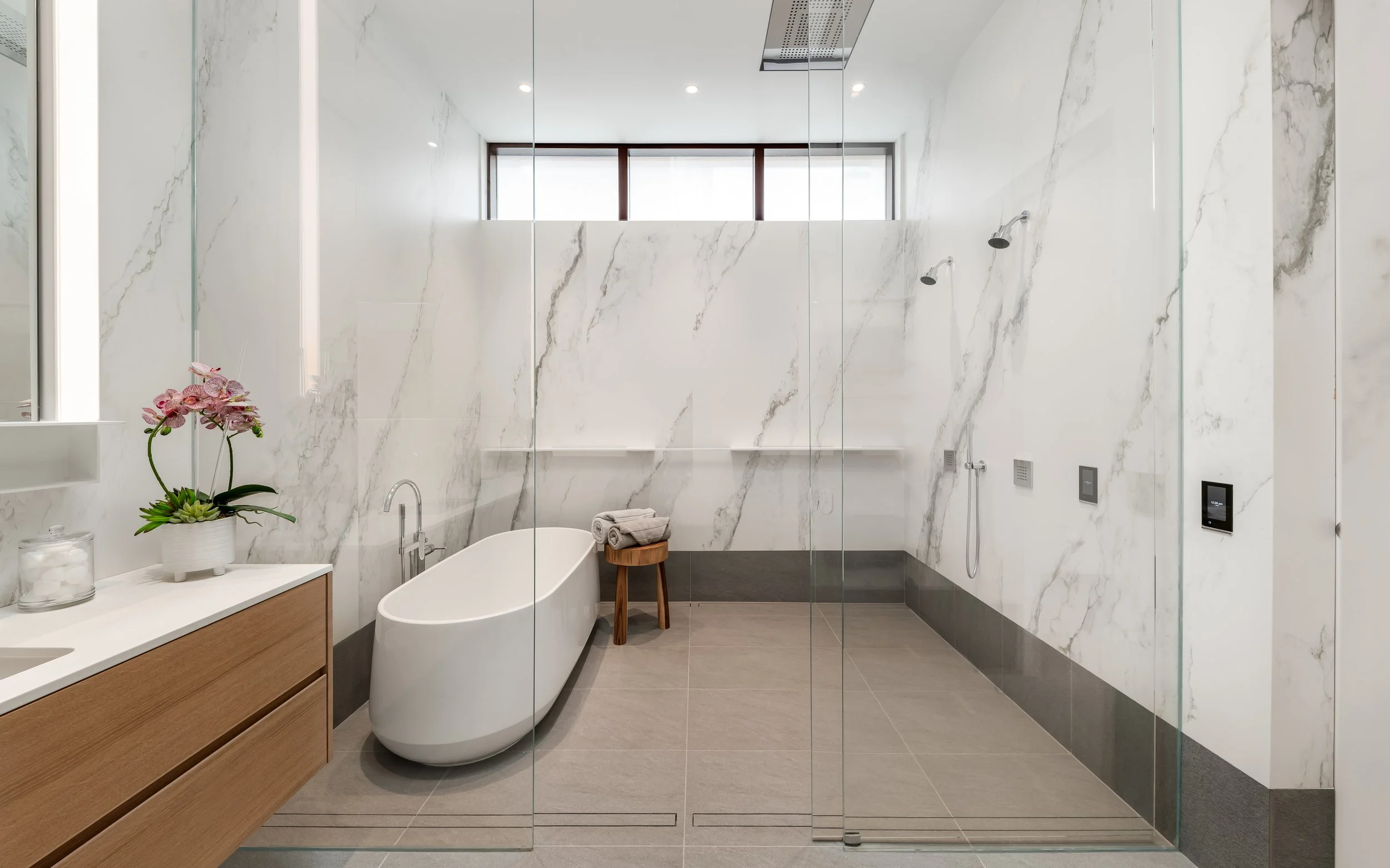 Modern bathroom with marble walls, a freestanding bathtub, a wooden stool with towels, and a glass-enclosed shower area.