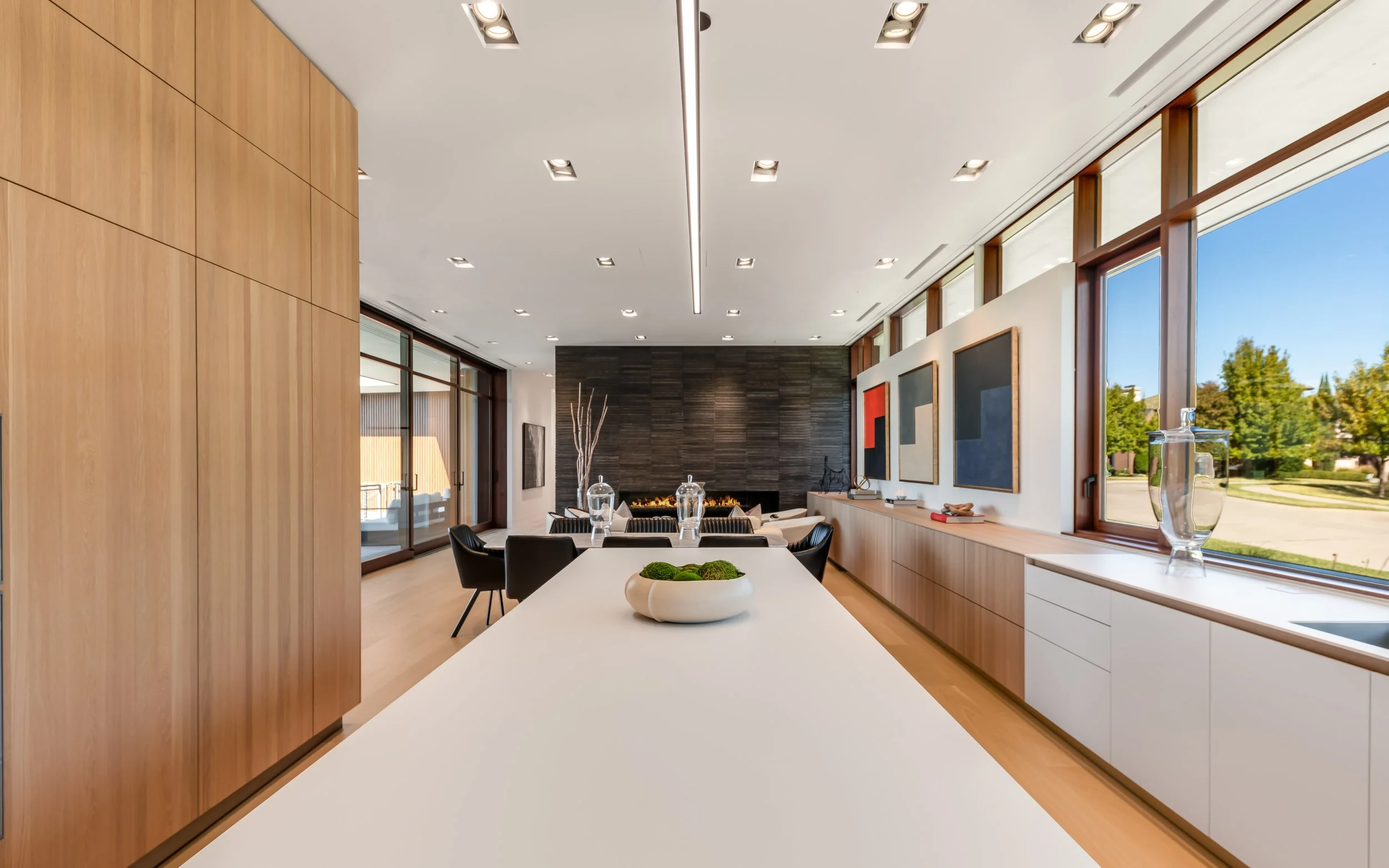 Modern dining area in a house with a large white island, black chairs, artwork on the walls, and a fireplace in the background, with large windows showing a green outdoor scene.