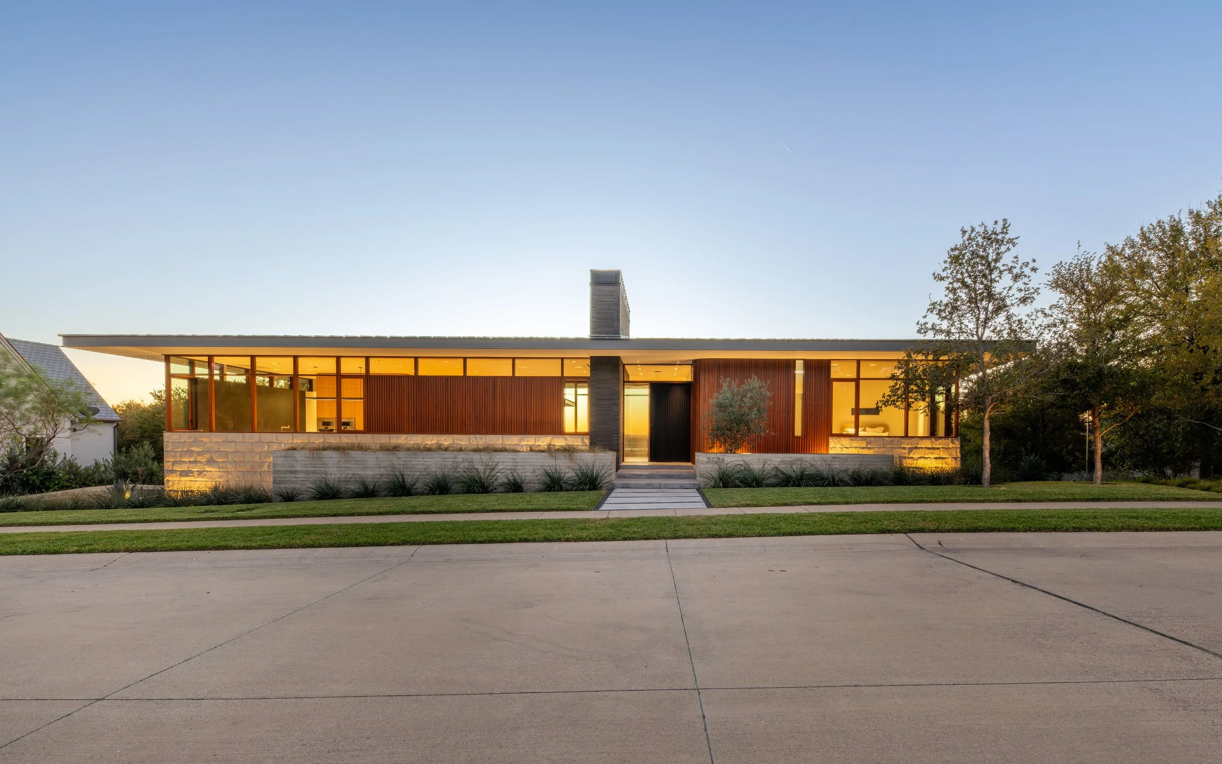 Modern single-story house with large glass windows, wooden paneling, a stone foundation, and a front walkway, set in a landscaped yard during sunset.