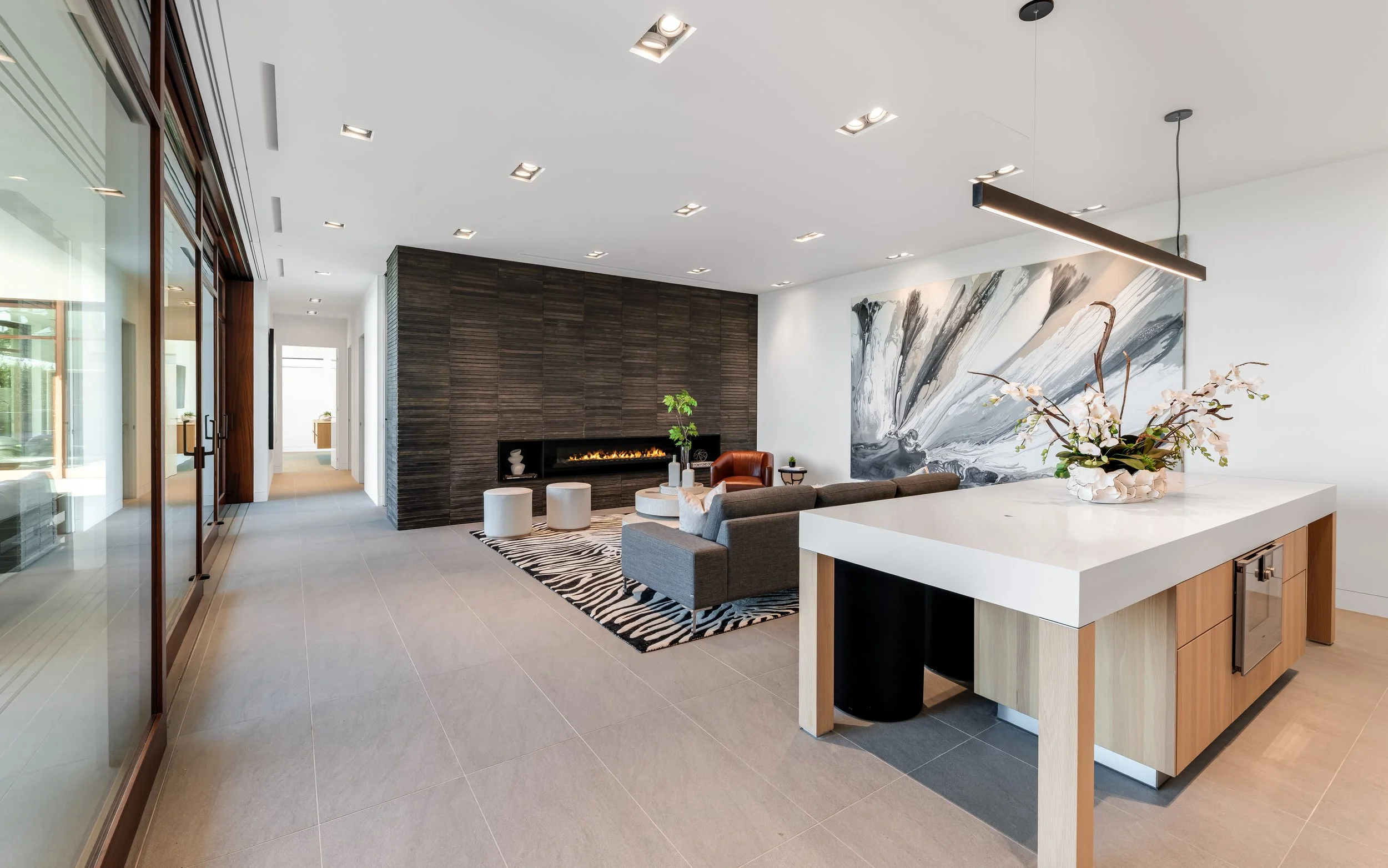 Modern living room with gray sectional sofa, leather armchair, fireplace, large abstract wall art, and a white island with a flower arrangement.