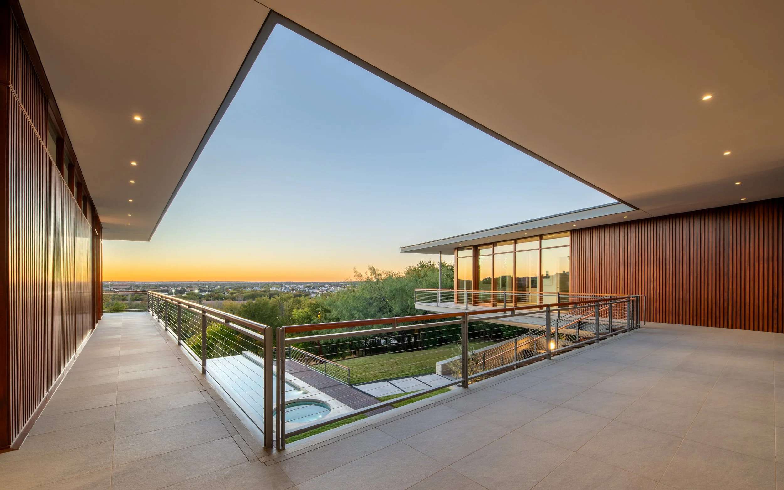 Modern residential balcony with wood-paneled walls, glass railing, tiled flooring, overlooking a view of trees, a swimming pool, and a city skyline at sunset.