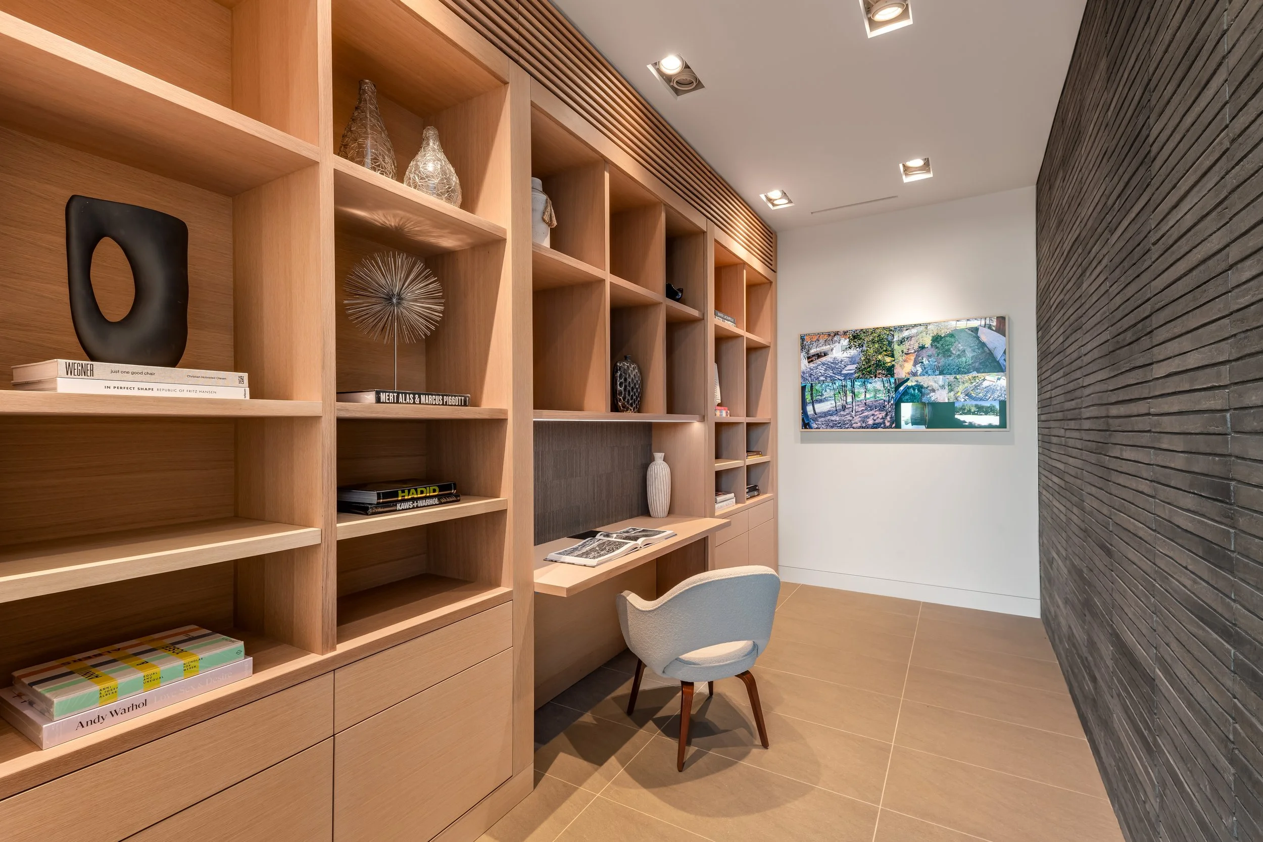 Interior of a modern office or library with wooden shelves filled with decorative items and books, a small desk with a chair, and a wall-mounted digital screen displaying outdoor scenery.