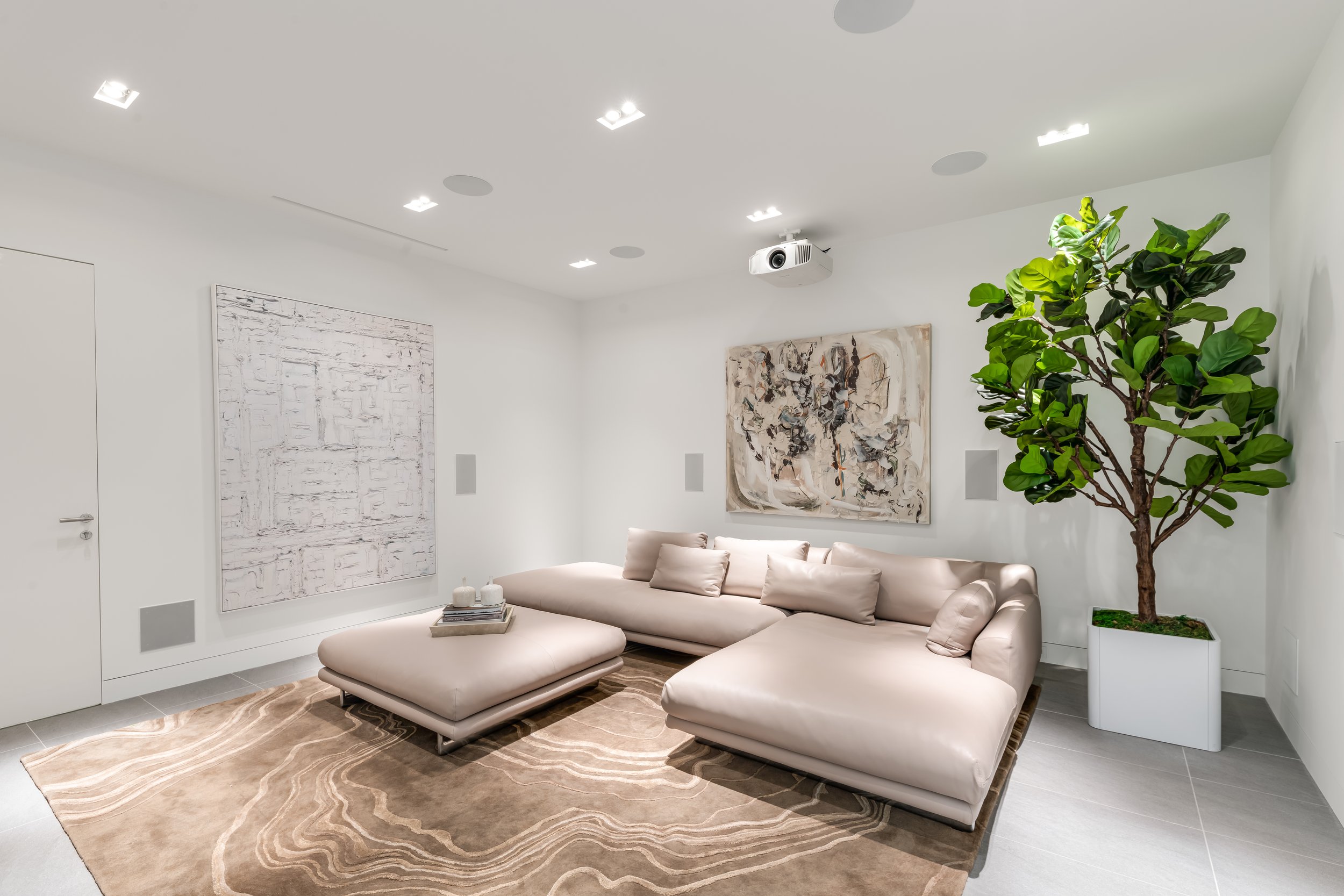 Modern living room with white walls, a beige sectional sofa with matching pillows, a large abstract painting, a potted plant, a textured area rug, and a ceiling projector.