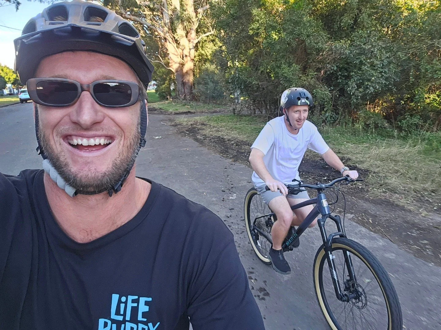 Two men riding bikes outdoors on a trail, both wearing helmets and sunglasses, with trees and grass in the background.