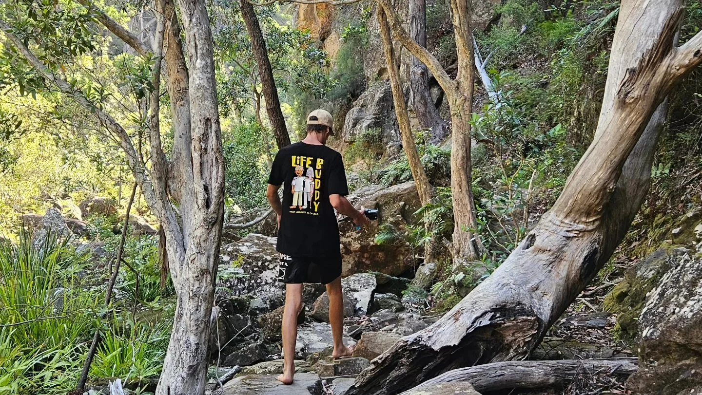 A person with long hair, wearing a black t-shirt and shorts, walking barefoot on a rocky trail in a forest, holding a camera.