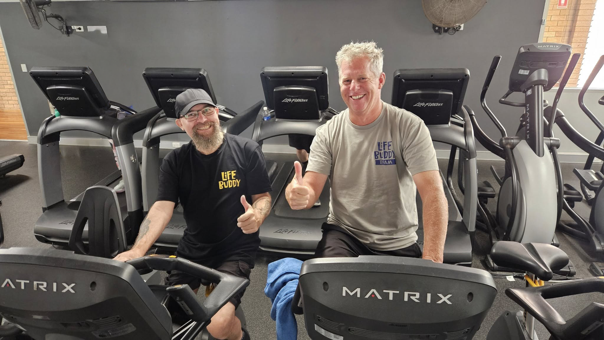 Two men smiling and giving thumbs up while sitting on exercise machines in a gym, wearing matching t-shirts that say 'LIFE BUDDY'. Behind them are additional exercise equipment and a gray wall.
