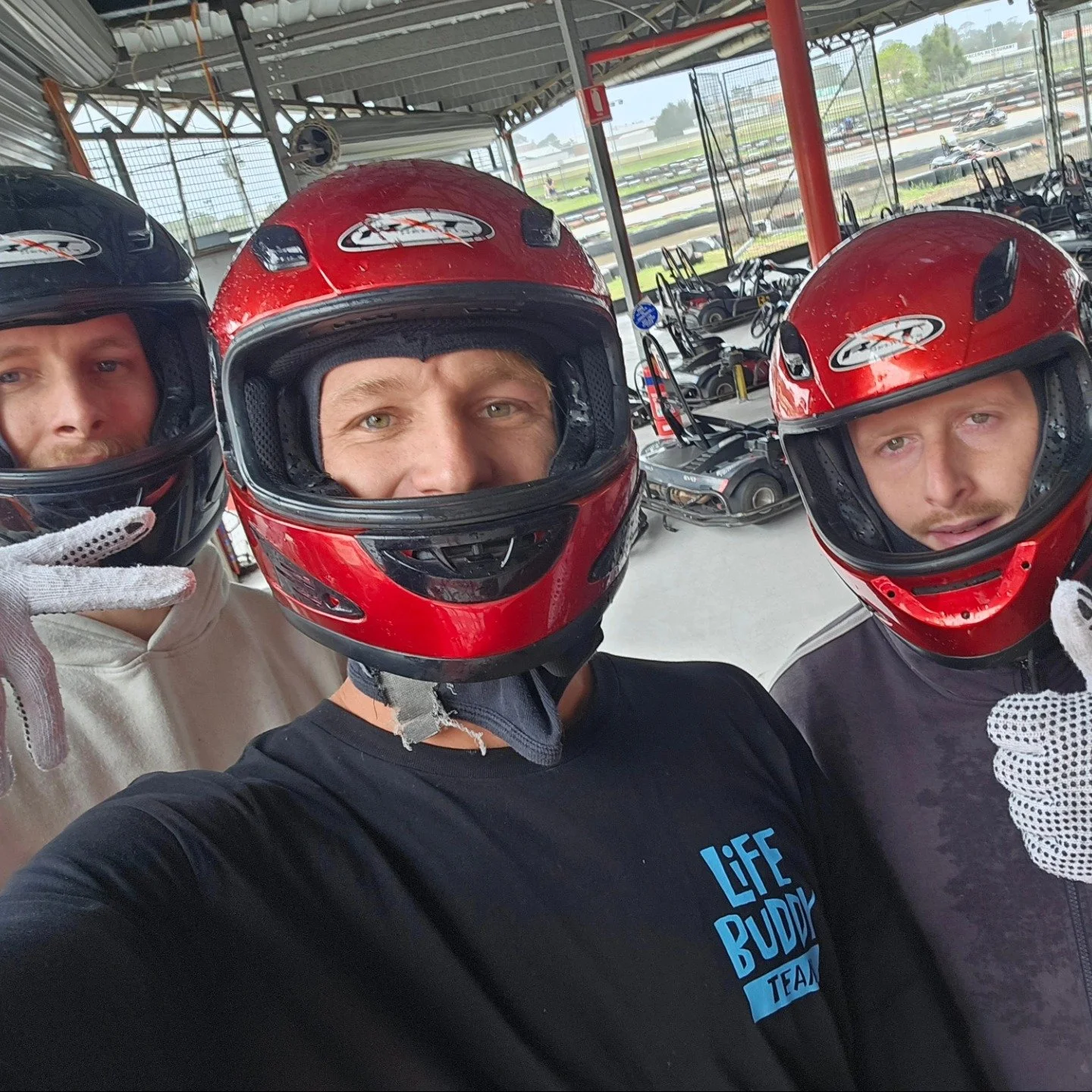 Three men wearing racing helmets and gloves taking a selfie at go-kart track. They are inside a shelter area with go-karts and the track visible in the background.