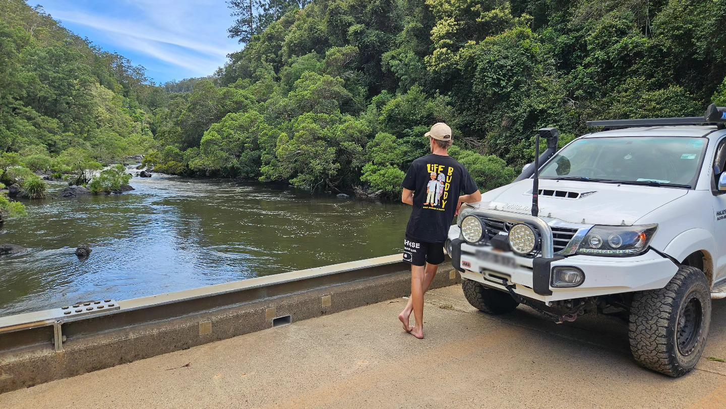 A man in a black t-shirt and shorts stands barefoot on a concrete roadside beside a white off-road vehicle, looking at a river surrounded by lush green trees and mountains under a partly cloudy sky.