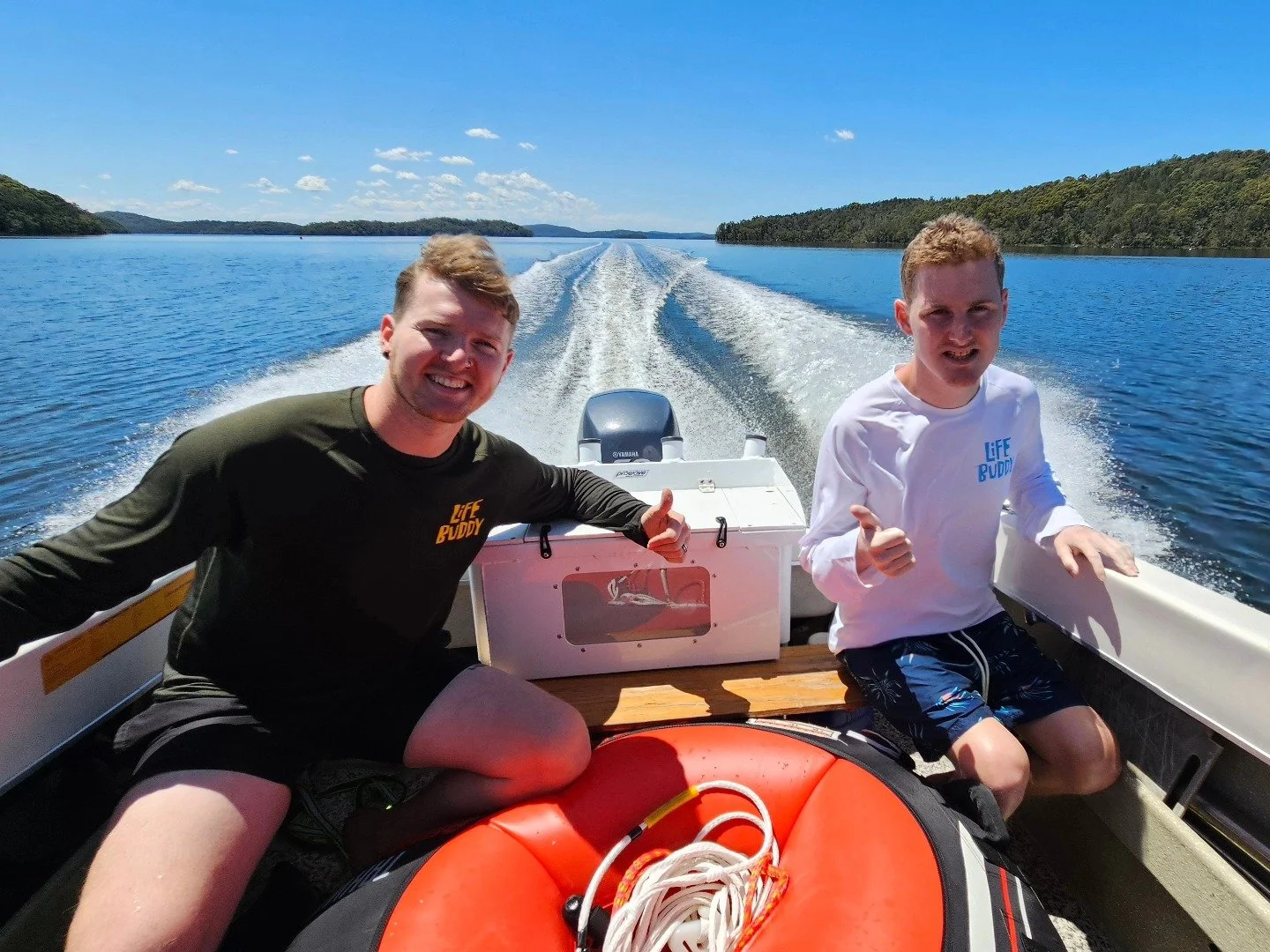 Two smiling men on a boat, giving thumbs up, with water and clear sky in the background.