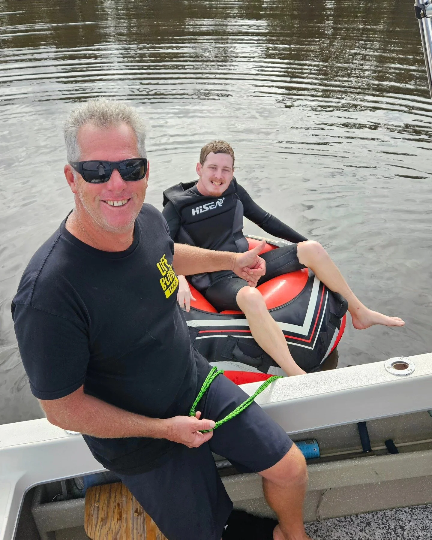 A man and a young man smiling on a boat, with the young man sitting in a water tube on the water, giving a thumbs-up sign, while the man holds a green rope attached to the water tube.