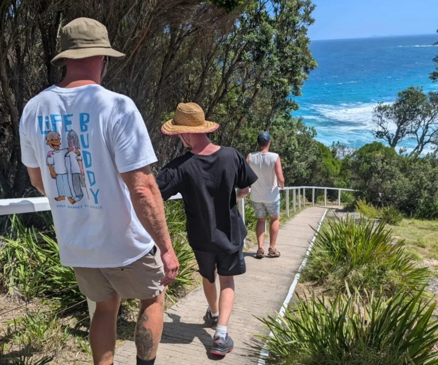 Three people walking along a coastal trail with ocean view, surrounded by trees and greenery on a sunny day.