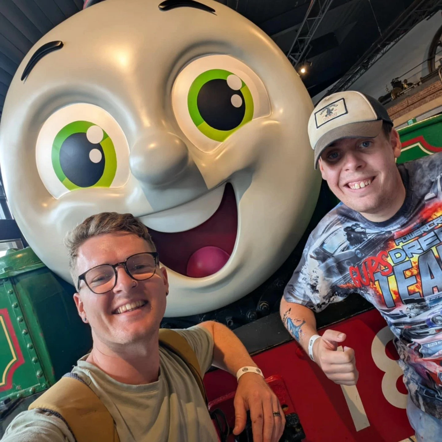 Two young men taking a selfie with a large smiling cartoon character that has big green eyes and a big open mouth, in an amusement park setting.