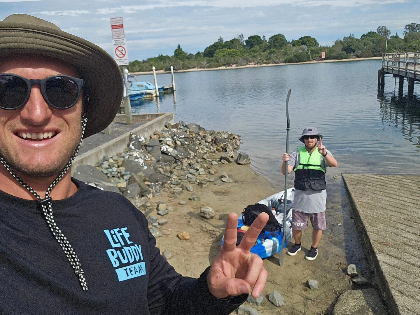 Two men at a shoreline near water, one in the foreground taking a selfie with sunglasses and a hat, and the other in the background holding a kayak paddle and wearing a life jacket, with a kayak and gear beside him.