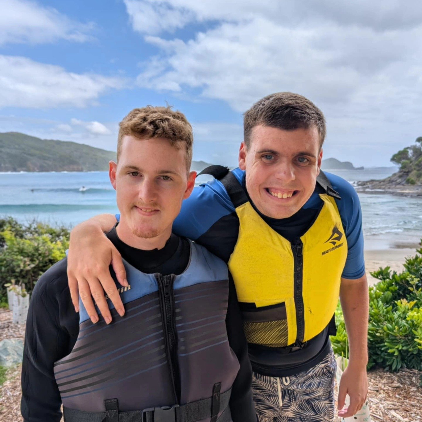 Two smiling young men standing outdoors near the beach with arms around each other's shoulders, wearing life jackets, with ocean, small islands, and a partly cloudy sky in the background.