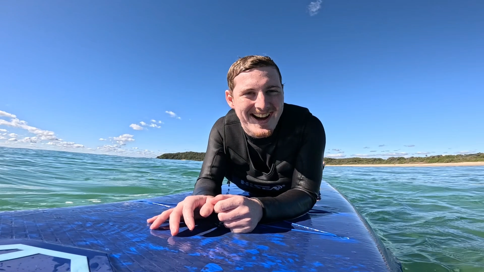 Young man smiling on a surfboard in the ocean on a clear day with a blue sky and distant shoreline.