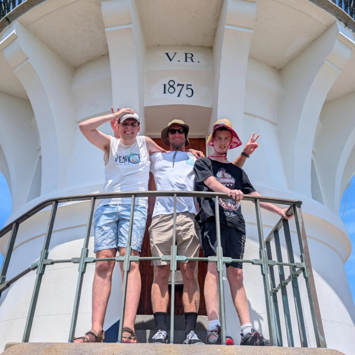 Three people standing on a balcony of a white lighthouse, smiling and posing for a photo, with a blue sky background. The lighthouse has the date 1875 and the initials V.R.