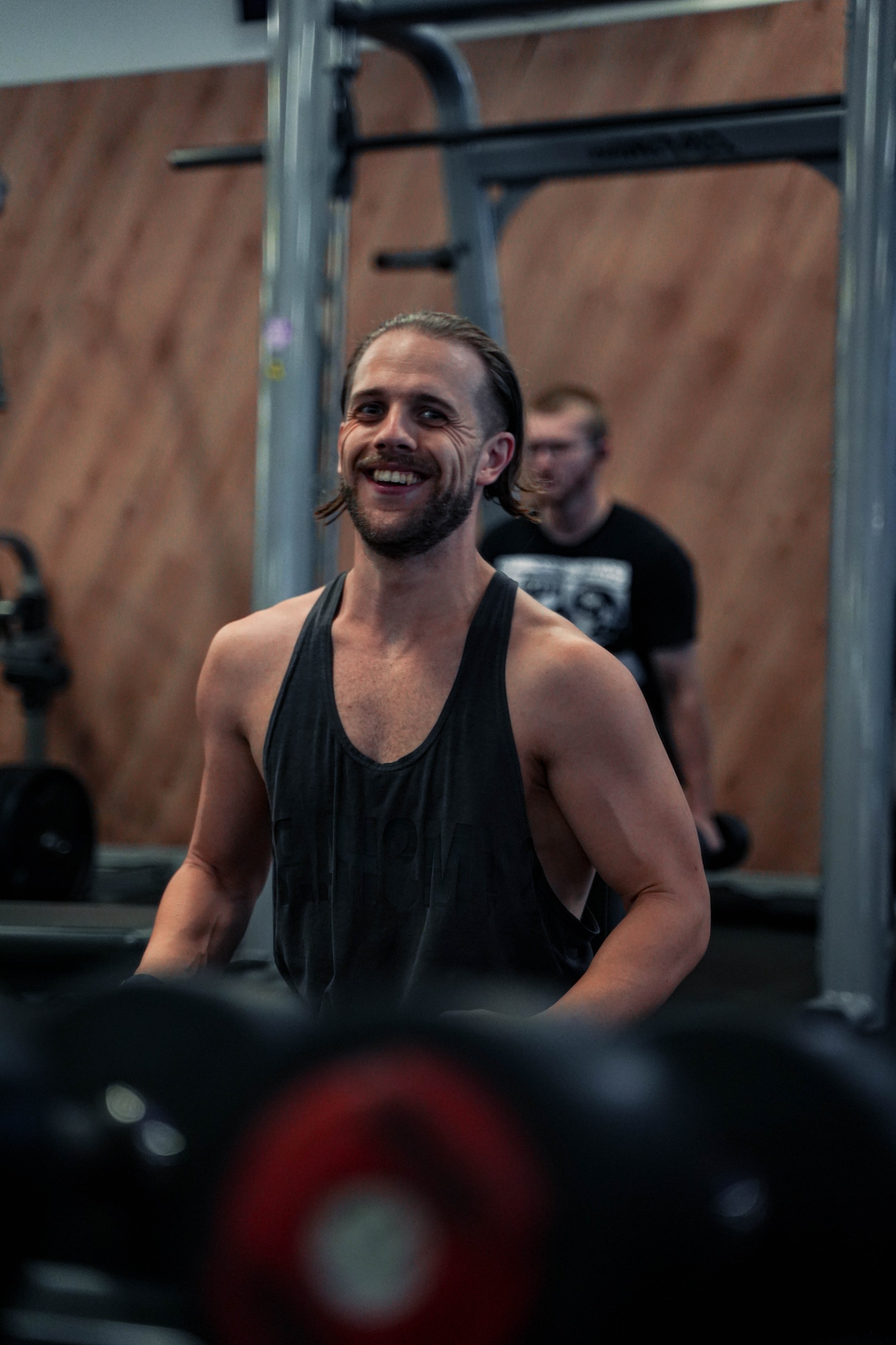 A smiling man in a black tank top lifting weights in a gym with a man in black t-shirt in the background.