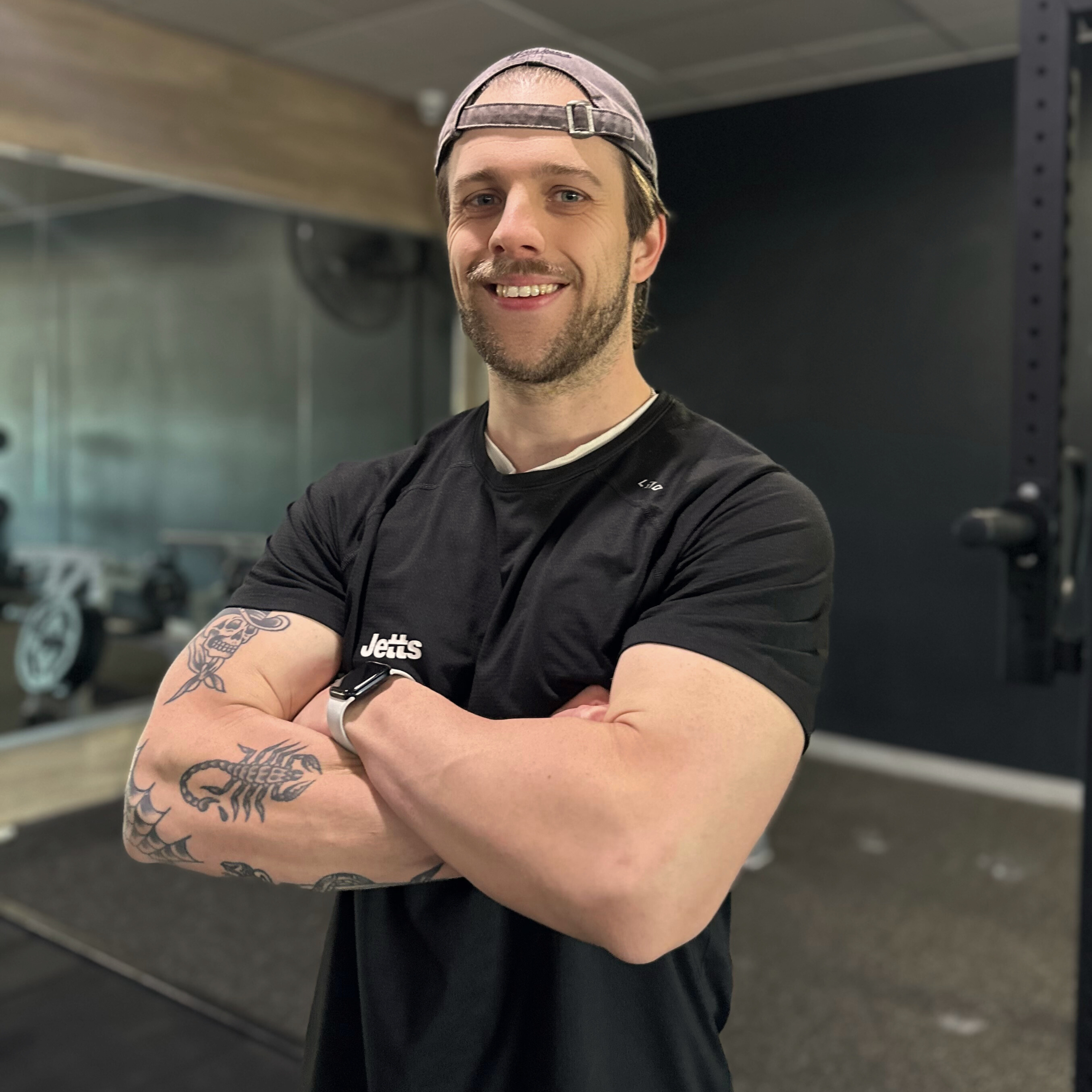 A young man with a beard, tattoos on his arm, wearing a black t-shirt with the word 'Jett's' on it, and a backwards gray cap, smiling with his arms crossed inside a gym.