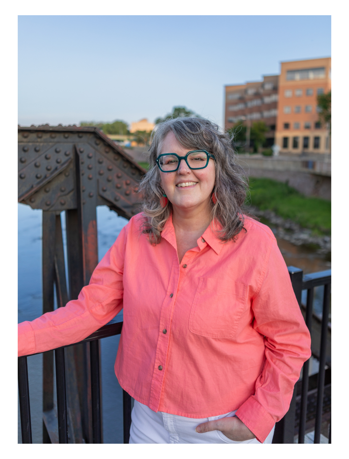 Woman with gray curly hair, wearing teal glasses, a coral pink blouse, and white pants, standing on a bridge by a river with buildings in the background during daytime.