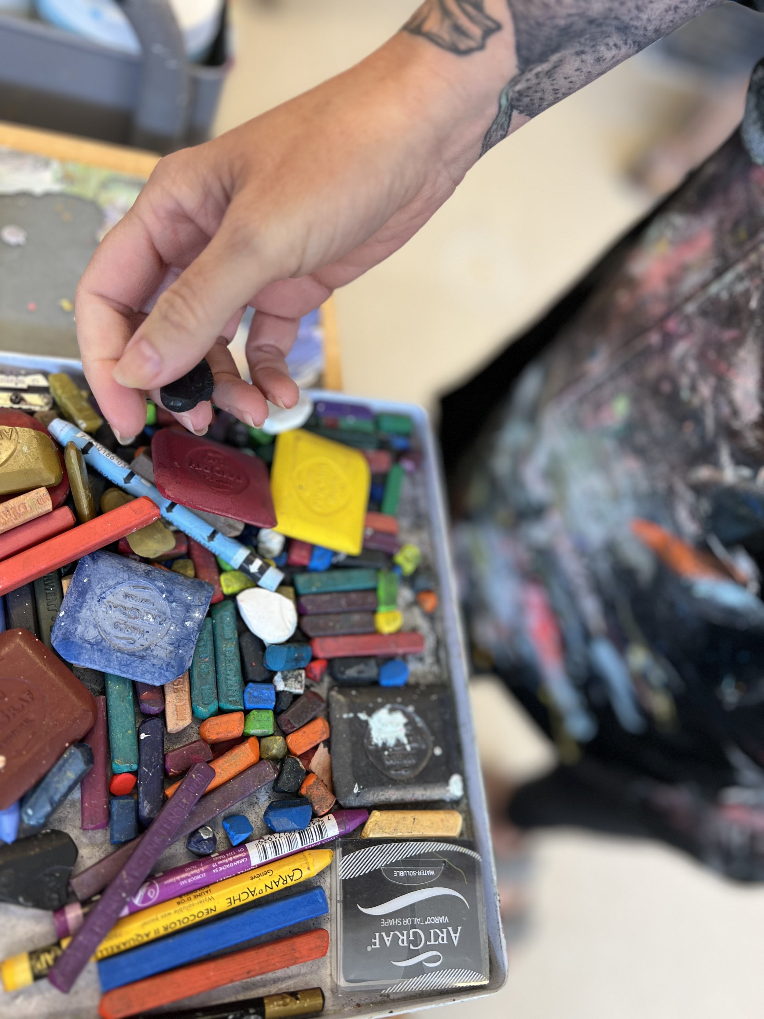 Close-up of a person's hand selecting a black pastel from a box filled with various colorful pastels and chalks, with a blurred artist's apron or smock in the background.