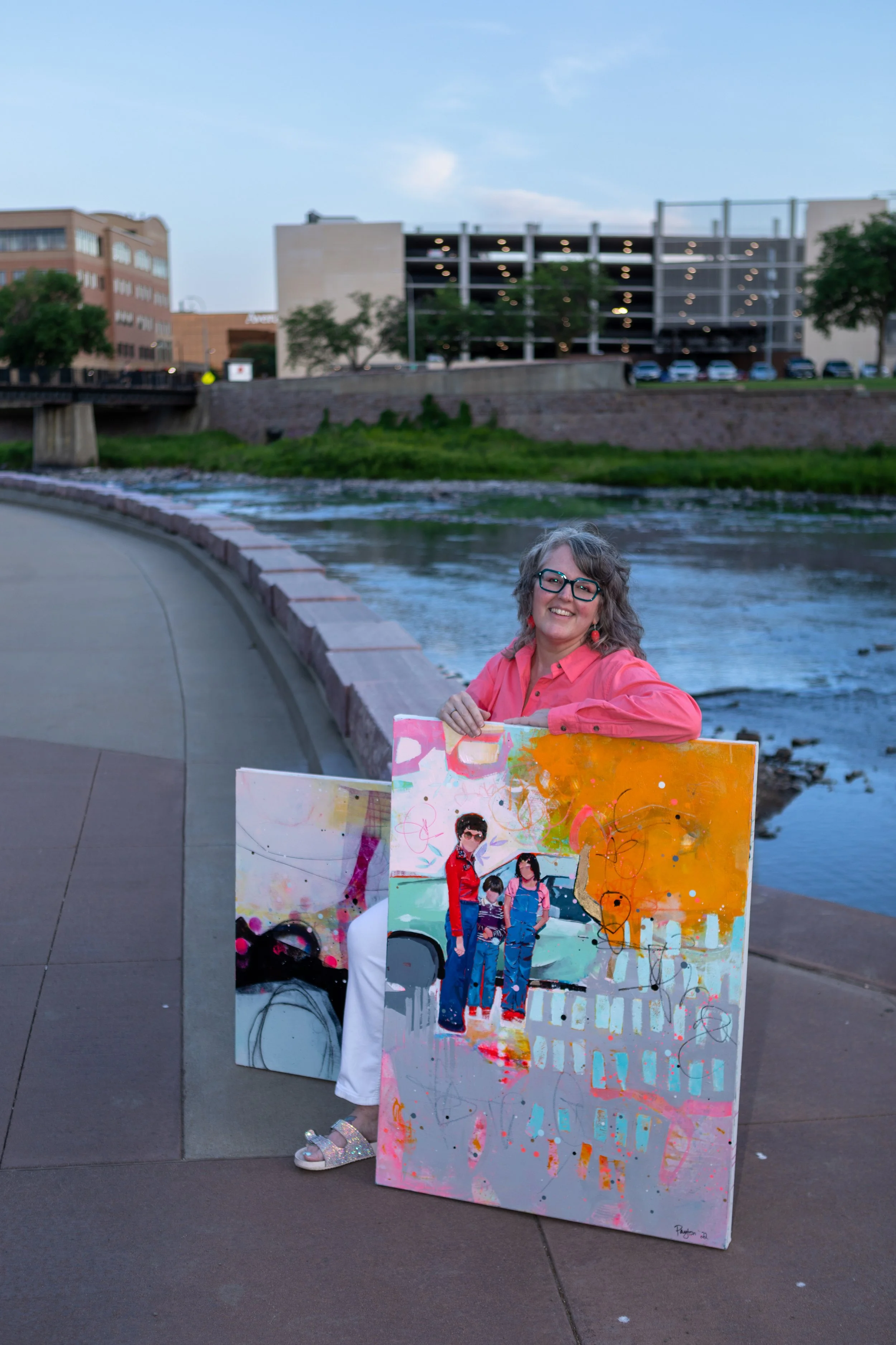 A woman in a pink shirt sitting next to her colorful abstract painting by a river with buildings in the background during twilight.