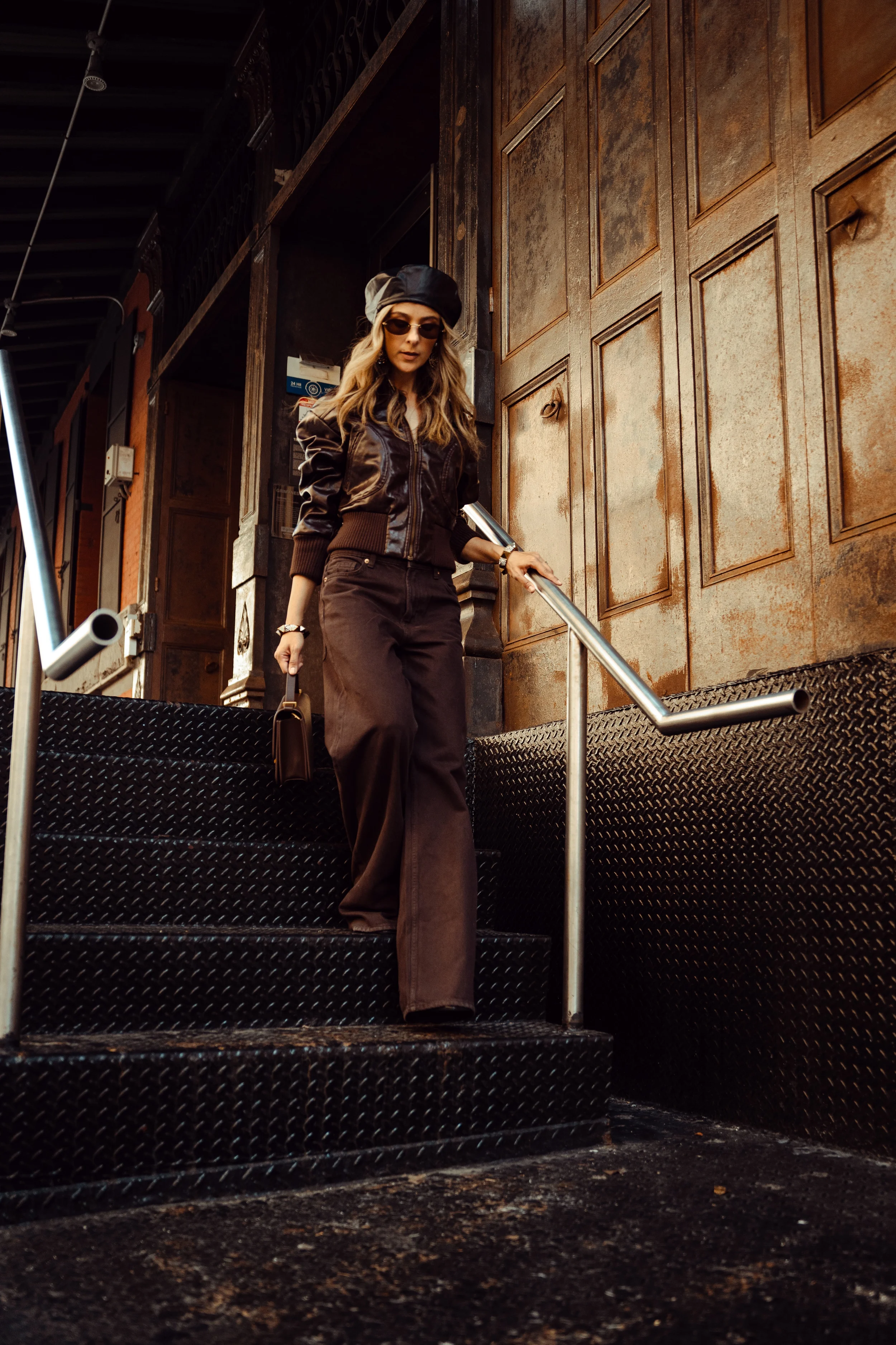 Alex in brown trousers, a matching jacket, sunglasses, and a beret walks down outdoor stairs next to a wall.