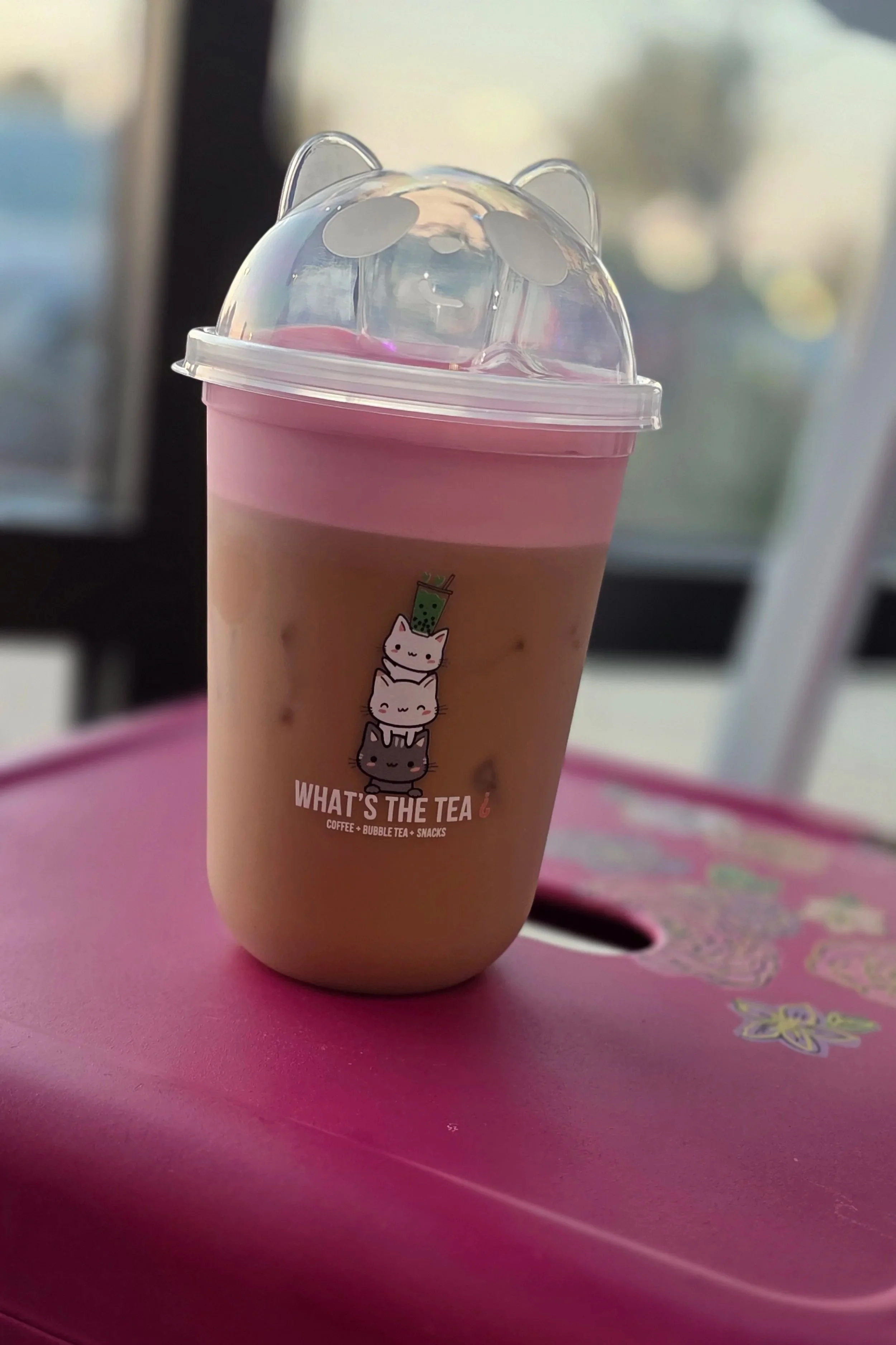 A cup of bubble tea with a pink and beige color, topped with a plastic lid that has bear ears. The cup features a logo with cartoon cats stacked vertically and the text 'WHAT'S THE TEA' underneath.