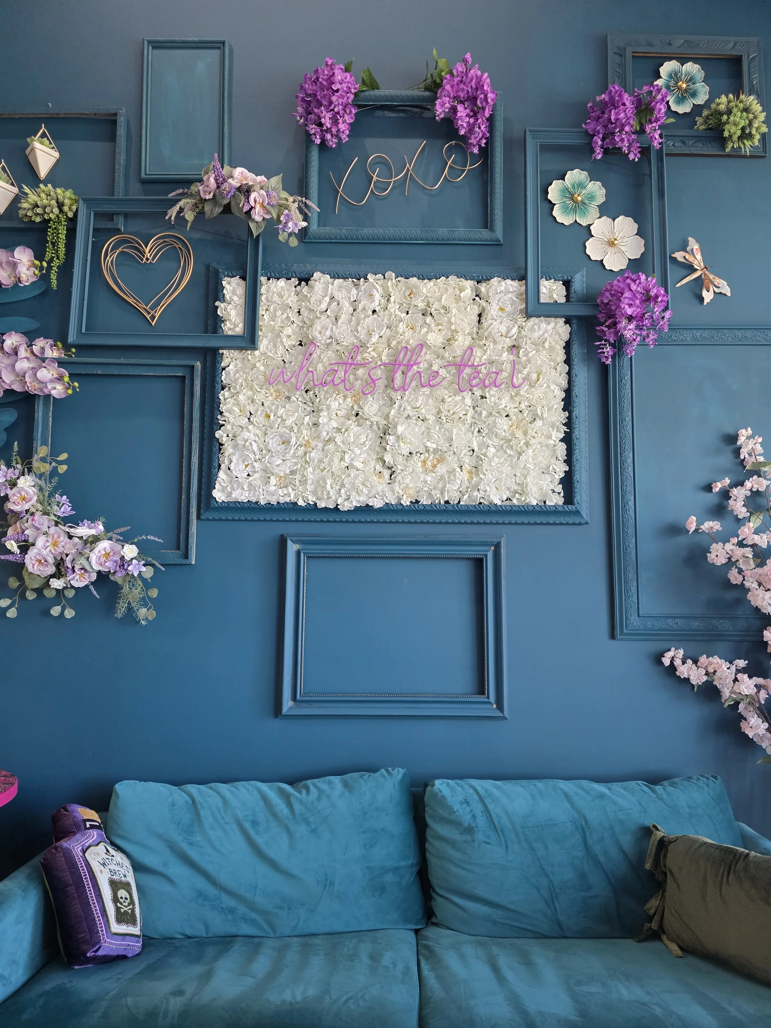 A decorated blue wall with empty picture frames, purple and white flowers, a pink neon sign that says 'what's the tea?', a white floral arrangement, and a blue sofa with purple and green pillows.