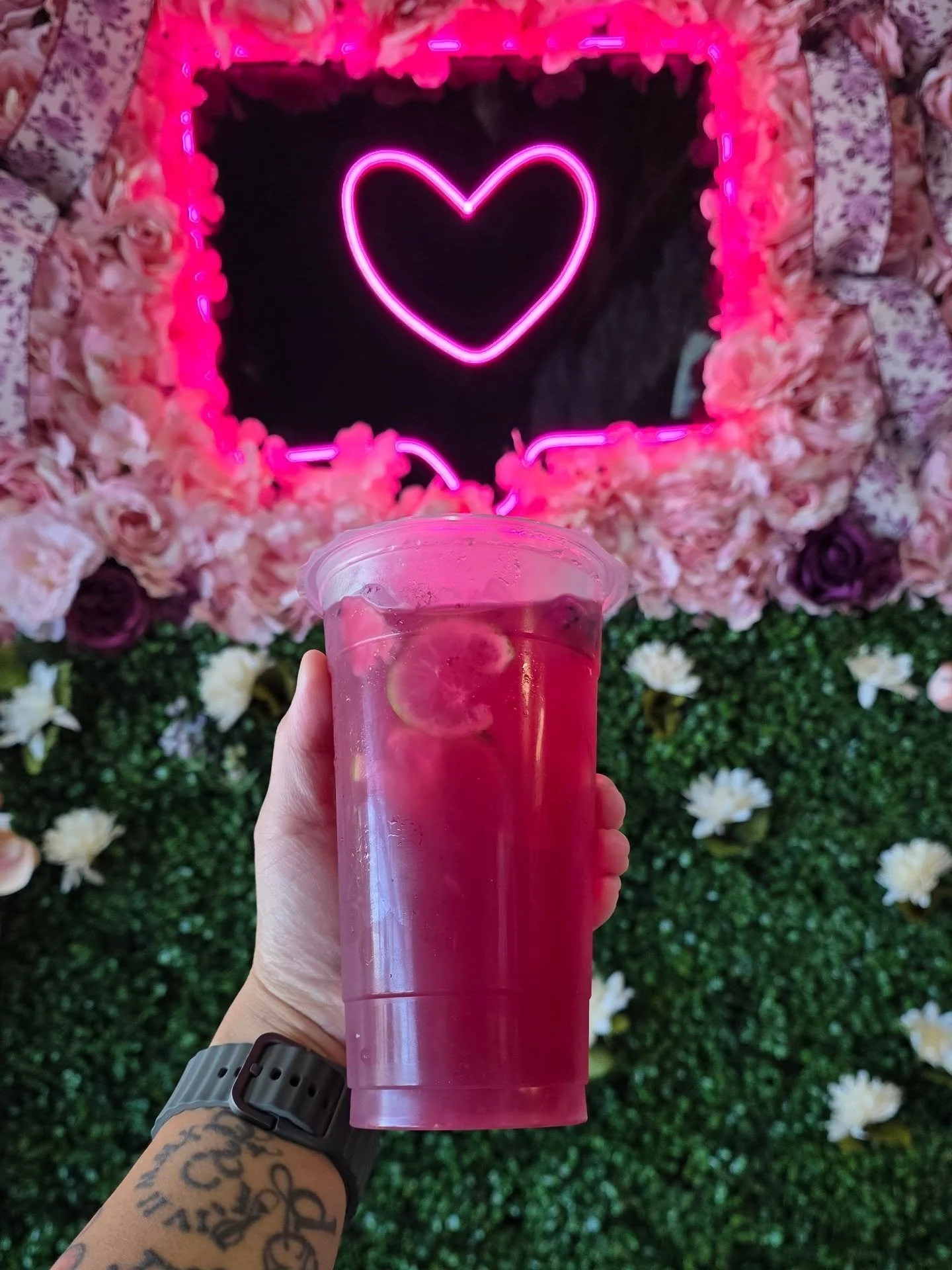 A hand holding a clear plastic cup with a pink drink and lemon slices in front of a decorative background featuring a neon pink heart and a border of pink and purple flowers.