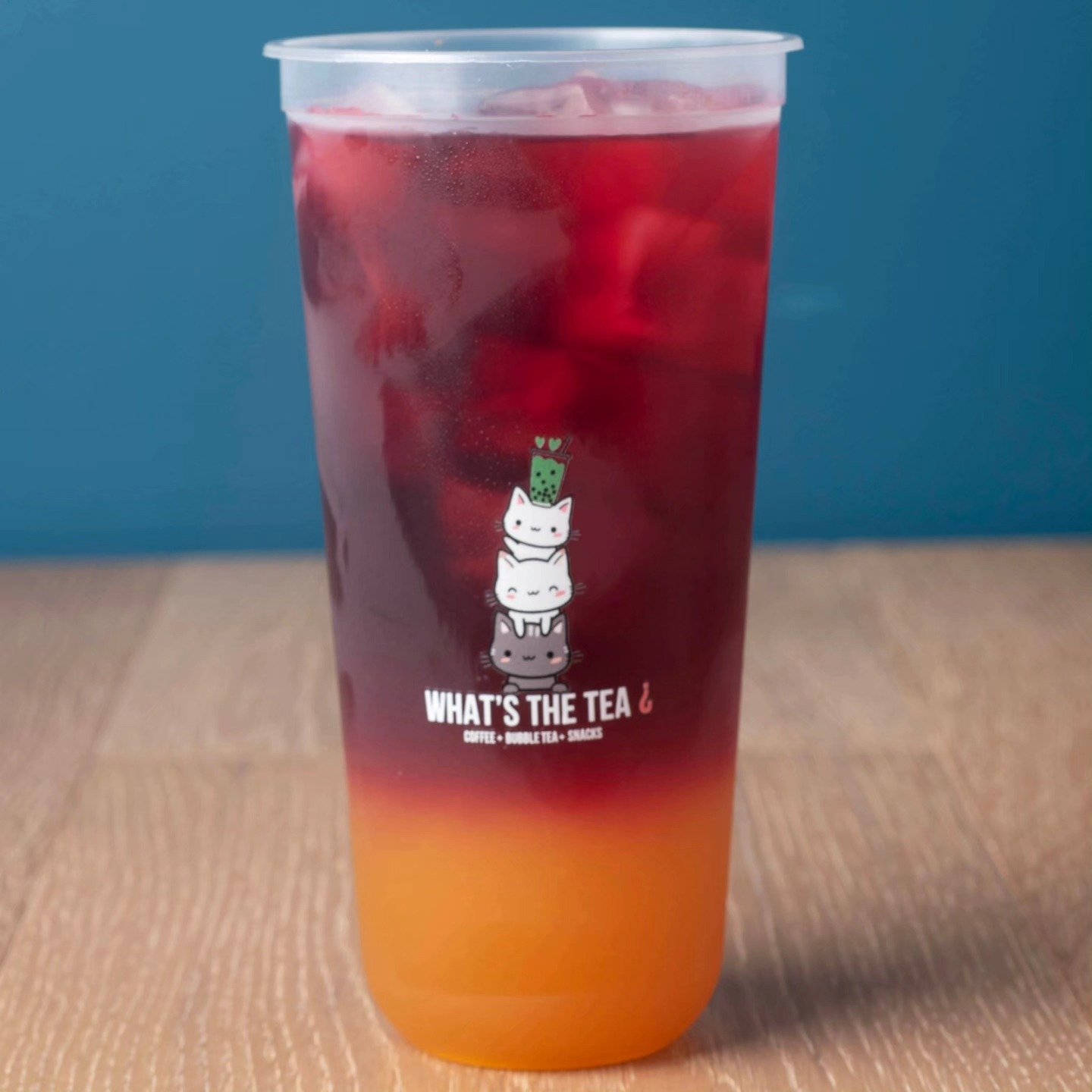 A tall plastic cup filled with a layered orange and red beverage with ice, featuring a sticker of cartoon cats stacked on top of each other with the text 'WHAT'S THE TEA' on the front.