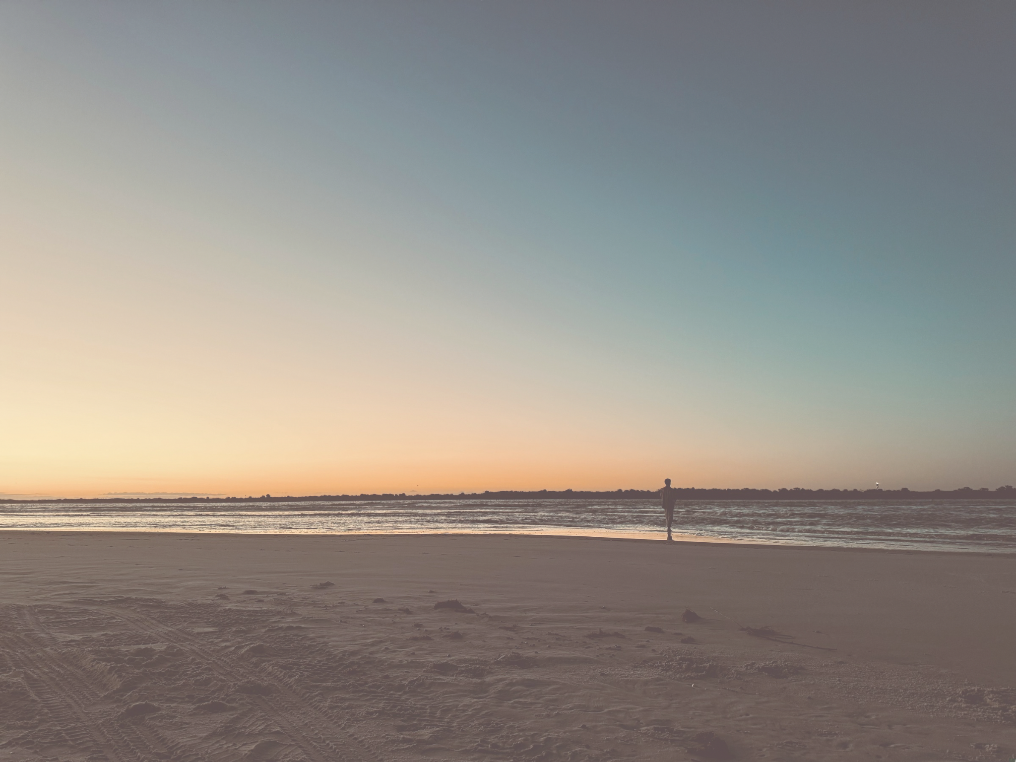 A lone person walking along the sandy beach near the water at sunset or sunrise, with a clear sky and calm ocean.