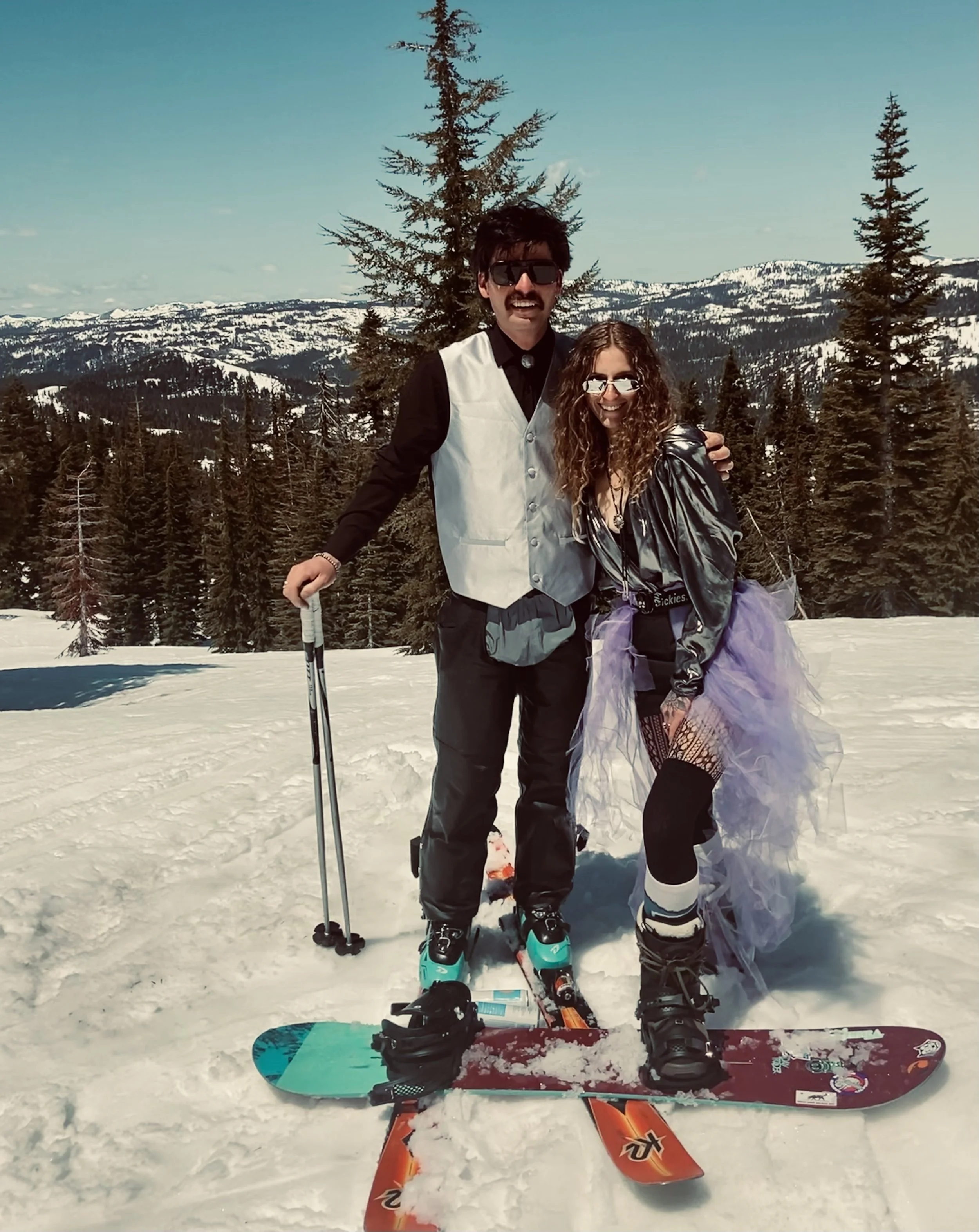 A man and woman are standing on snow-covered ground with skis and poles, smiling in front of a mountain landscape with trees and snow-capped peaks.