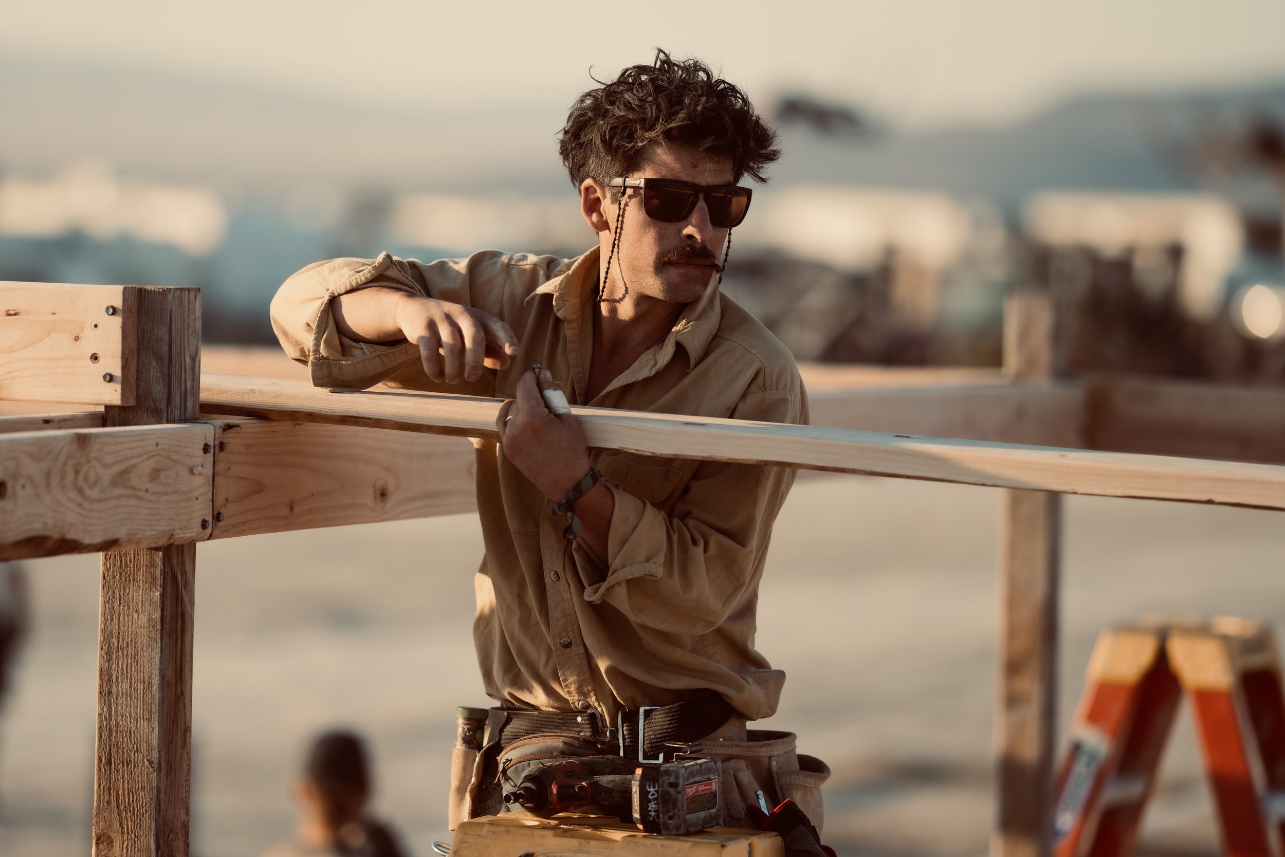 A man wearing sunglasses works on wooden construction outdoors near water.