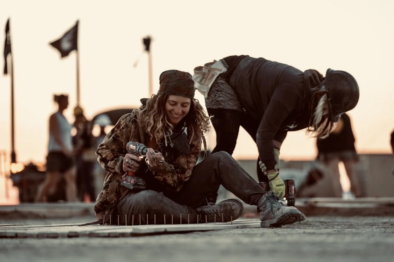 Two women working on a construction site at sunset, one sitting and holding a drill and the other bending down, both wearing work clothes and safety gear.