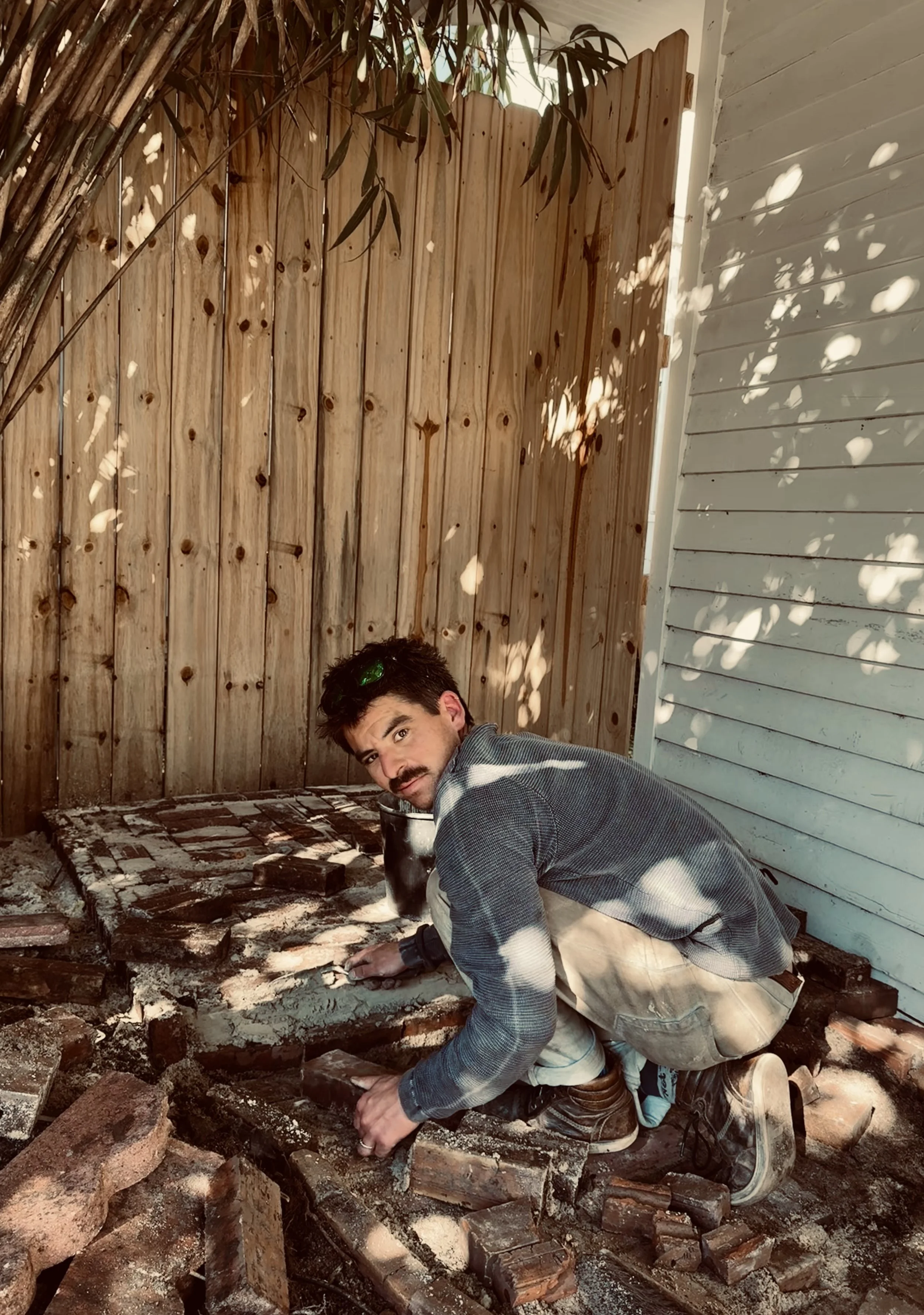 A man crouching on his knees working on a brick floor in a backyard, with a wooden fence and house siding visible in the background.