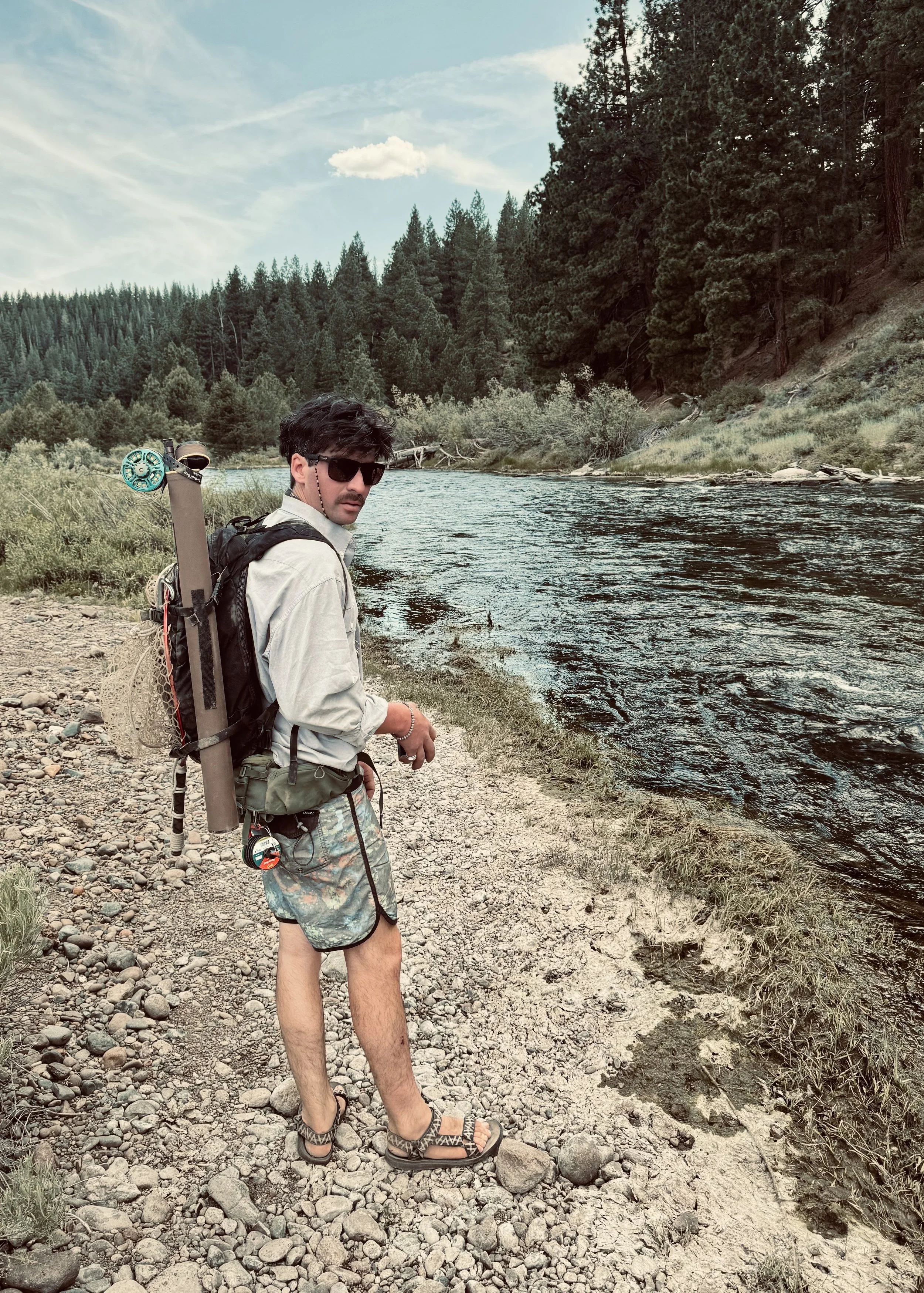 A man with sunglasses, a long-sleeve shirt, shorts, and sandals, standing on a rocky riverbank during daytime, with a backpack and a fishing rod attached to it, overlooking a river surrounded by trees and mountains.