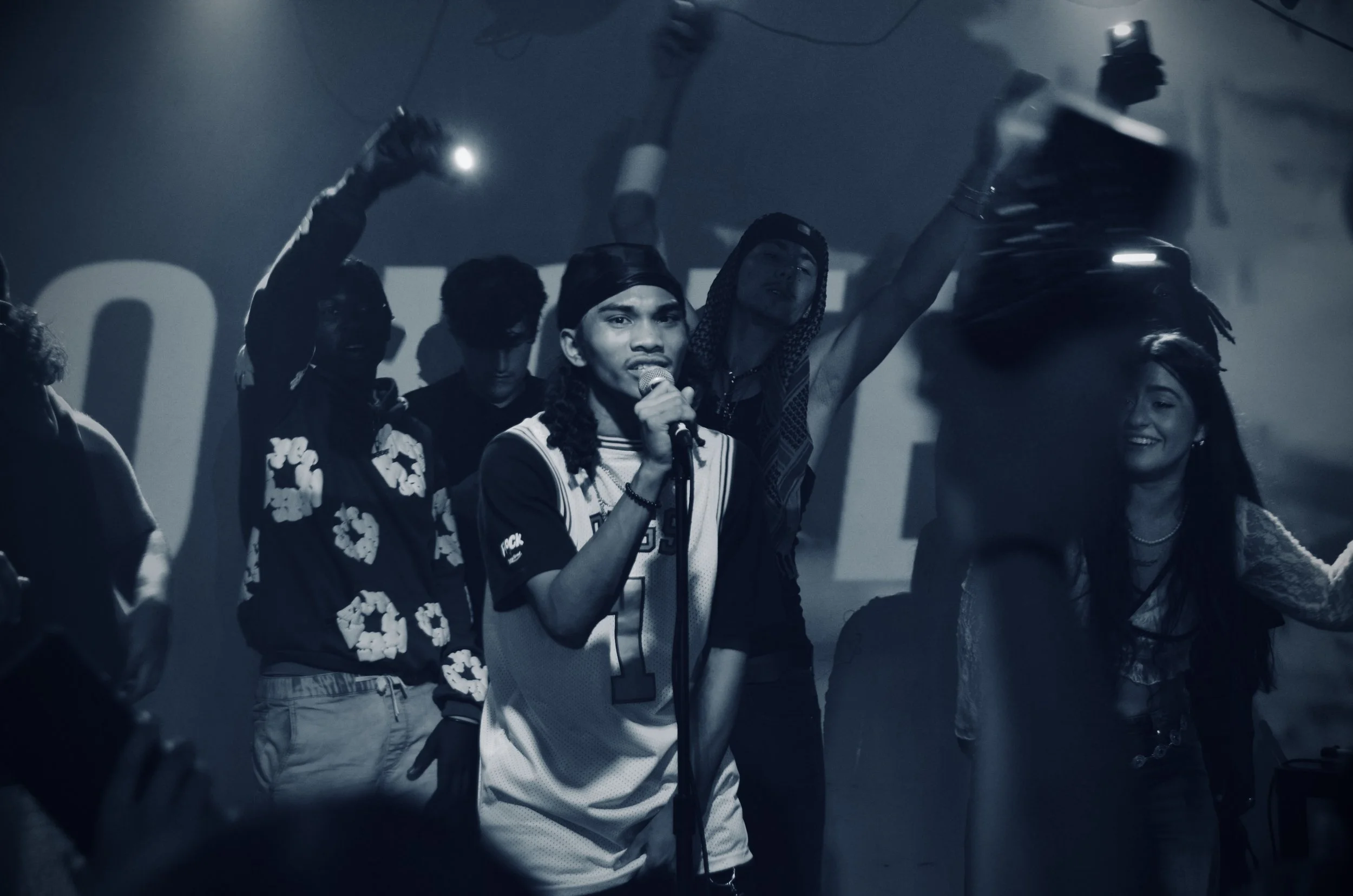 A group of young people on stage, with one person in the center holding a microphone and singing, surrounded by others raising their hands in the air, with a smoky background.