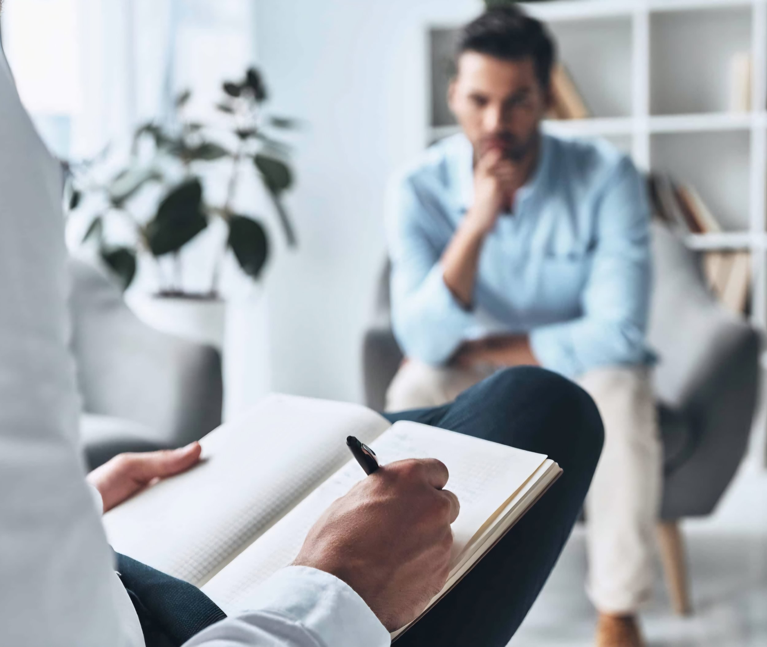A person holding a pen and writing in a notebook during a therapy or counseling session with a patient sitting across in a cozy, bright room.