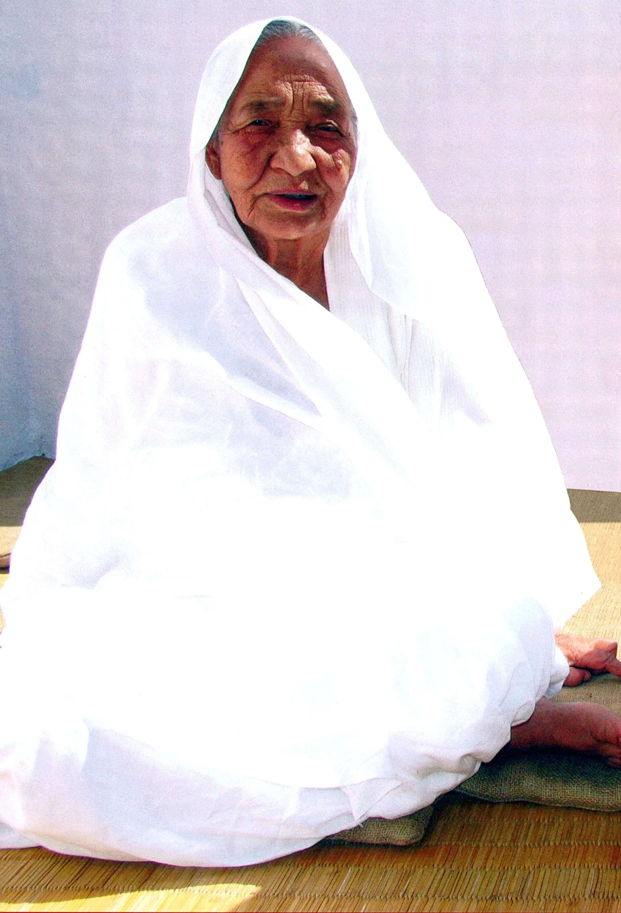 An elderly woman wearing a white traditional garment sitting on a mat, smiling.