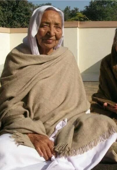 An elderly woman sitting outside in a beige shawl, with a white headscarf, smiling, on a sunny day.