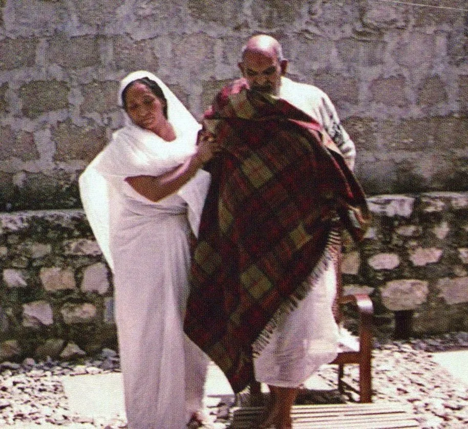 A woman in a white traditional dress and head covering appears to be touching or holding a man draped in a large colorful blanket, standing outside against a stone wall.