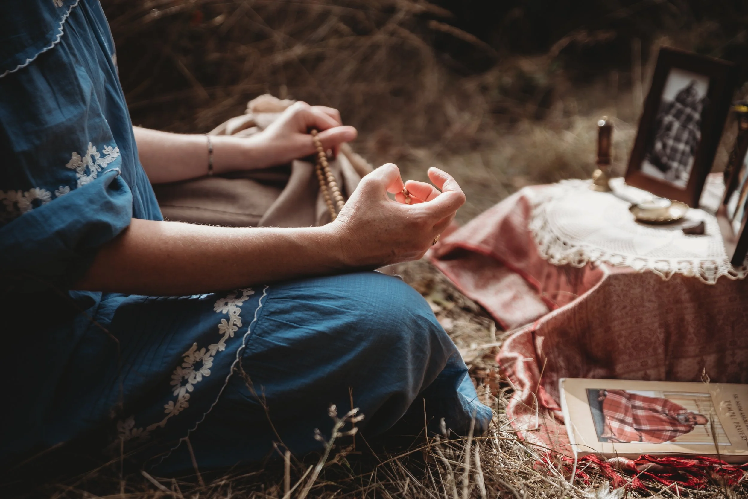 A woman sitting on the ground, holding a rosary, with a table beside her covered by a lace cloth, displaying framed photographs, a pocket watch, and other vintage items.