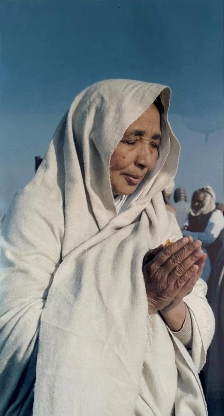 An elderly woman wearing a white shawl and holding her hands together in a prayer gesture, her eyes closed in a moment of devotion.