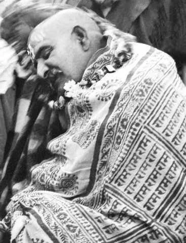 A man smiling and leaning on his hand, wrapped in a traditional patterned cloth with script, likely at a cultural or religious event.
