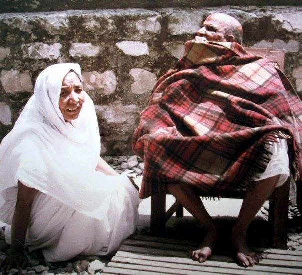 Elderly woman sitting on the ground smiling and talking to an elderly man wrapped in a plaid blanket, sitting on a stool against a stone wall.